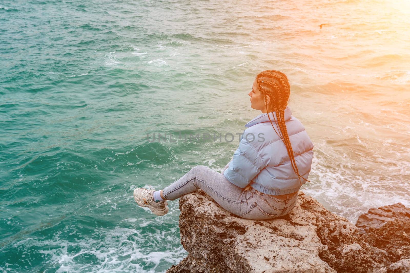 woman sea travel. A woman in a blue jacket sits on a rock above a cliff above the sea, looking at the stormy ocean. Girl traveler rests, thinks, dreams, enjoys nature by Matiunina