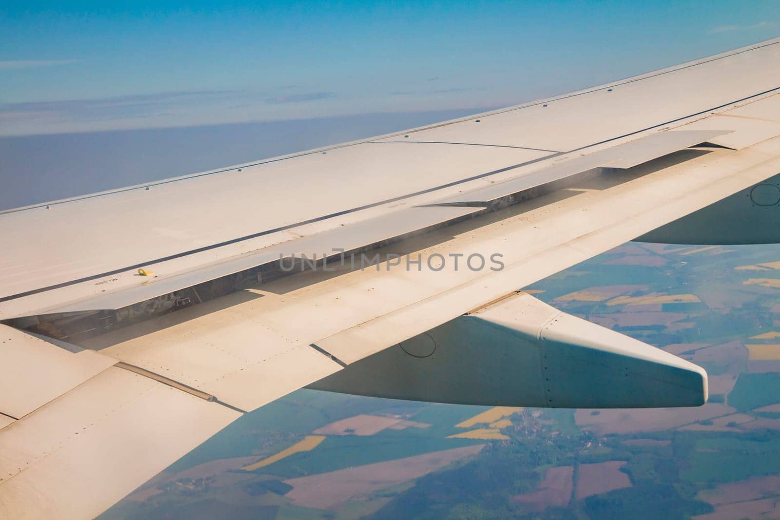 Airplane wing with the spoiler open on the sky over land.