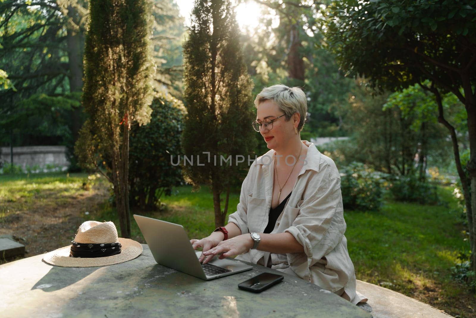 woman freelancer in glasses works at a computer at a white table in nature and spends her day productively by Matiunina