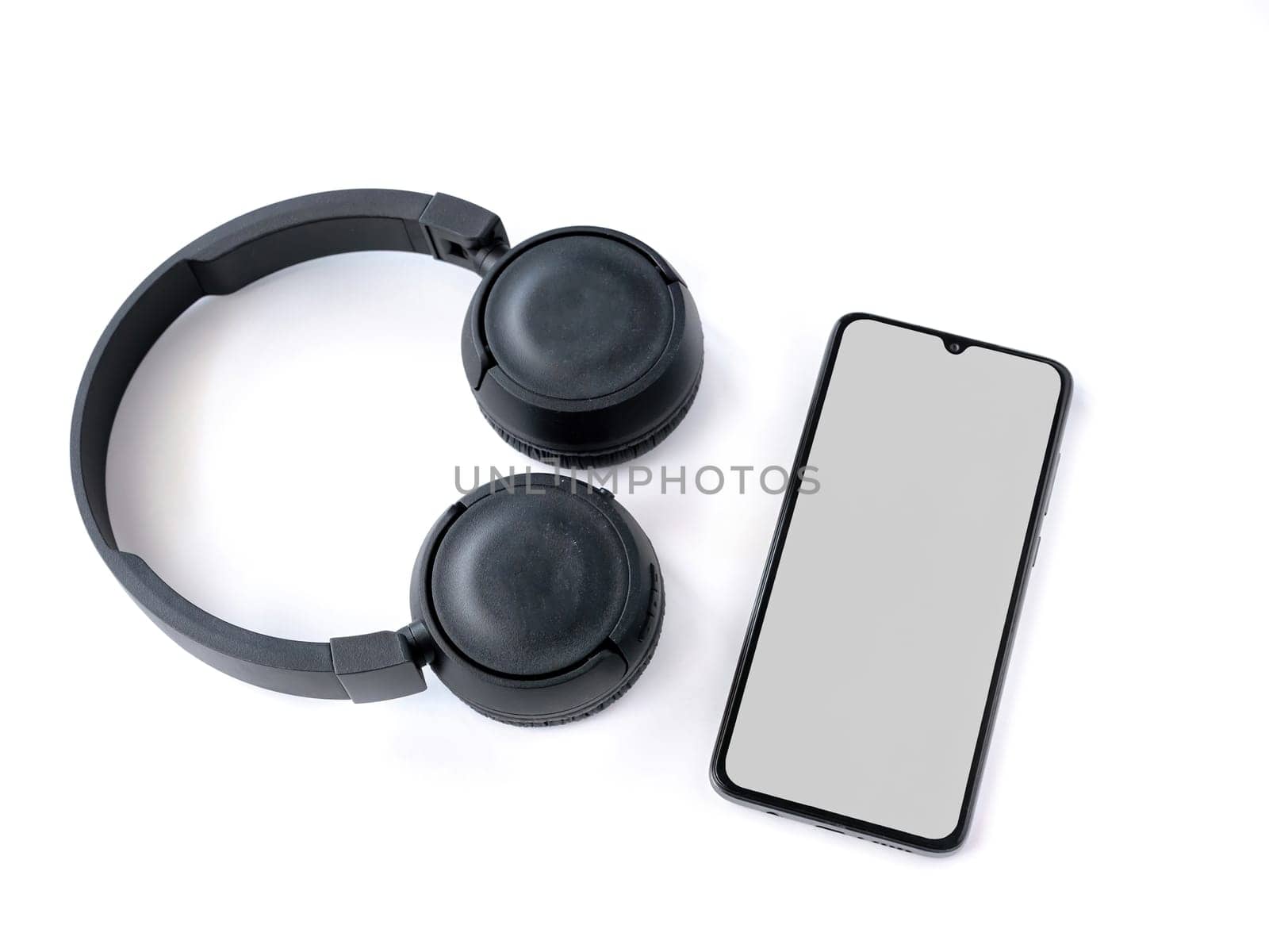 Black wireless headphone and mobile smartphone with a blank screen mockup lay on the surface of a white background. Top view flat lay with copy space. Music concept.