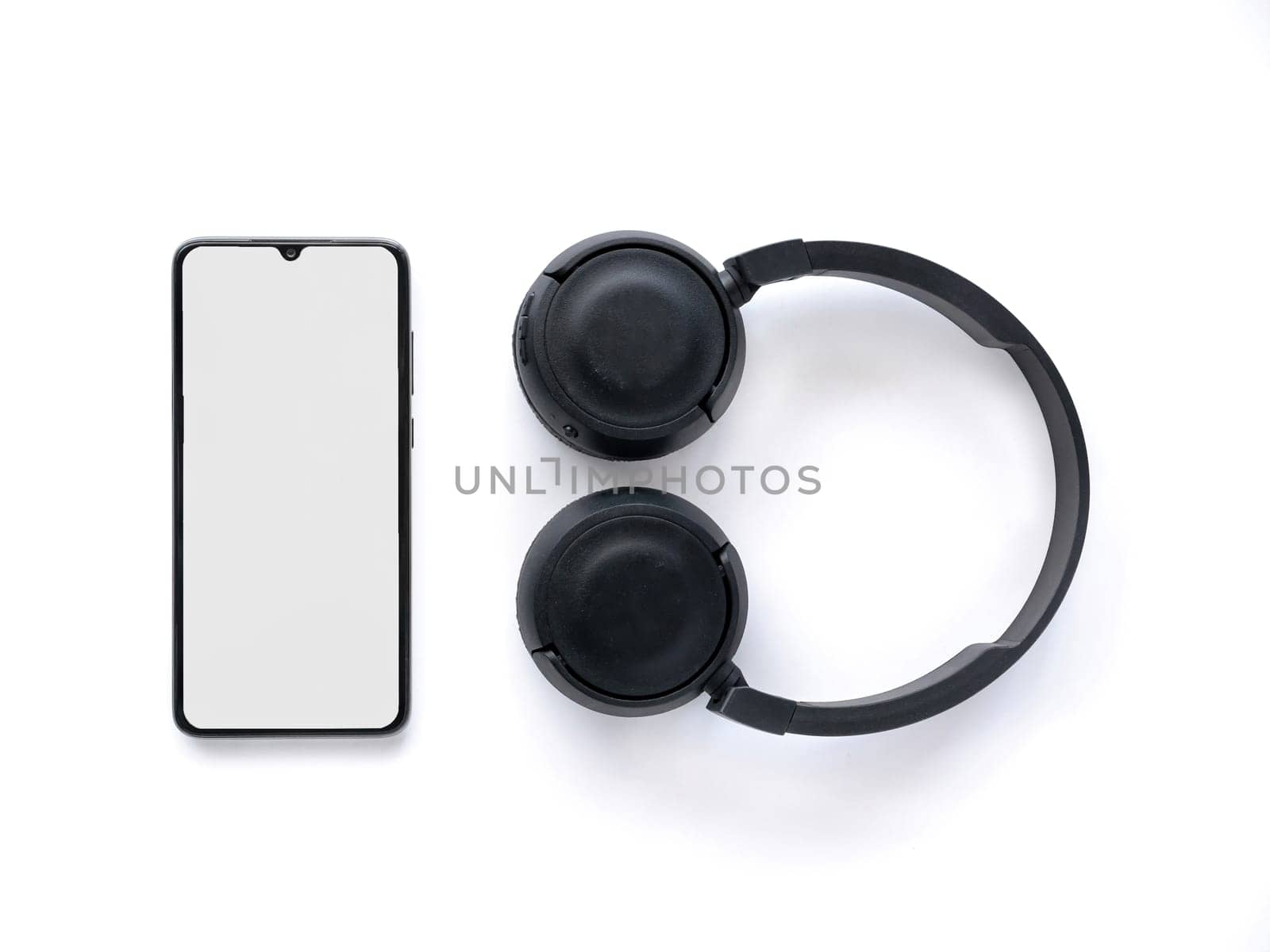 Black wireless headphone and mobile smartphone with a blank screen mockup lay on the surface of a white background. Top view flat lay with copy space. Music concept.