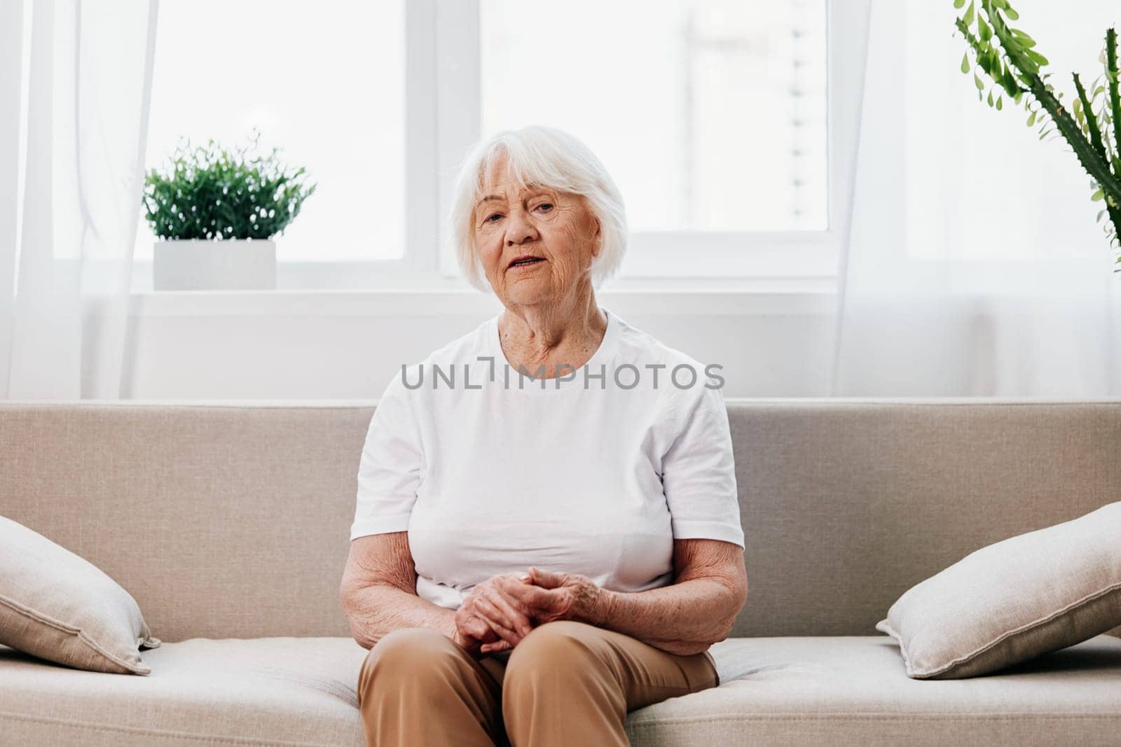 Elderly woman sits on sofa at home, bright spacious interior in old age smile, lifestyle. Grandmother with gray hair in a white T-shirt and beige trousers. by SHOTPRIME