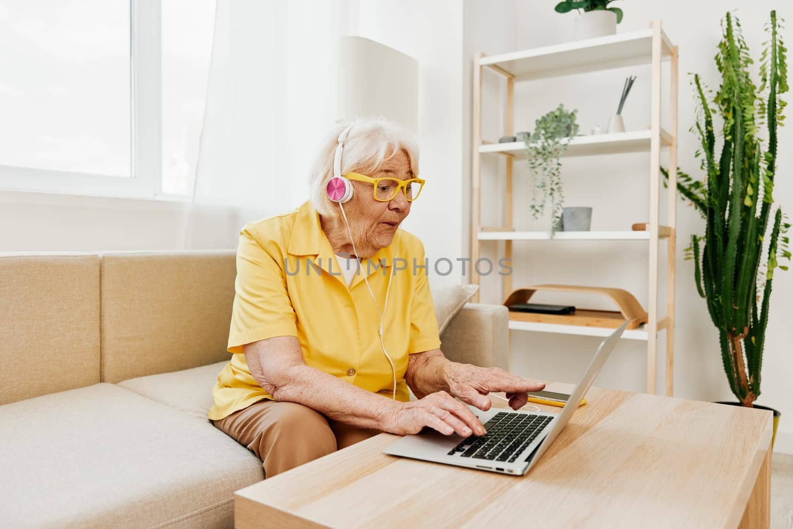 Happy elderly woman with a laptop typing with headphones sitting at home on the couch in a yellow shirt, bright modern interior, lifestyle online communication. High quality photo