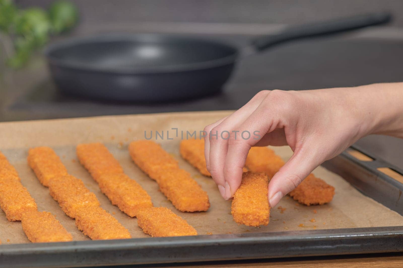 The hand lays out fish fingers against the background of a frying pan on special paper. The concept of frying fish sticks in the oven. Side view. by SERSOL