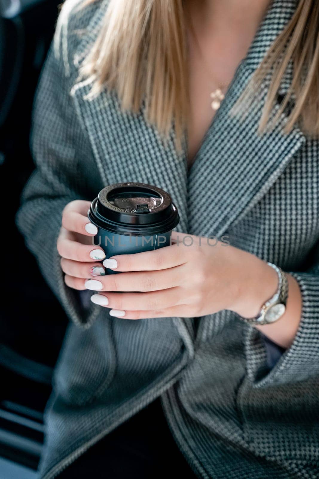 Happy woman coffee. she stands next to the car in the underground parking. Dressed in a gray coat, holding a glass of coffee in her hands, a black car