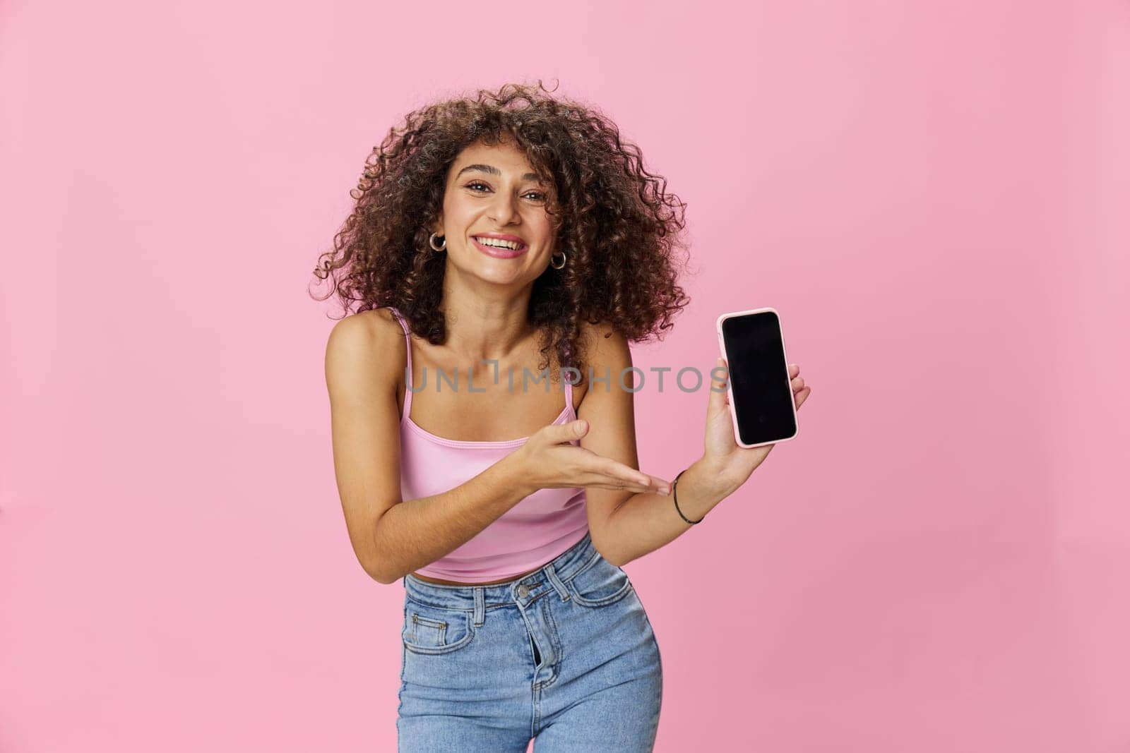 Woman blogger holding phone video call takes selfies, with curly hair in pink smile t-shirt and jeans poses on pink background, copy space, technology and social media, online by SHOTPRIME