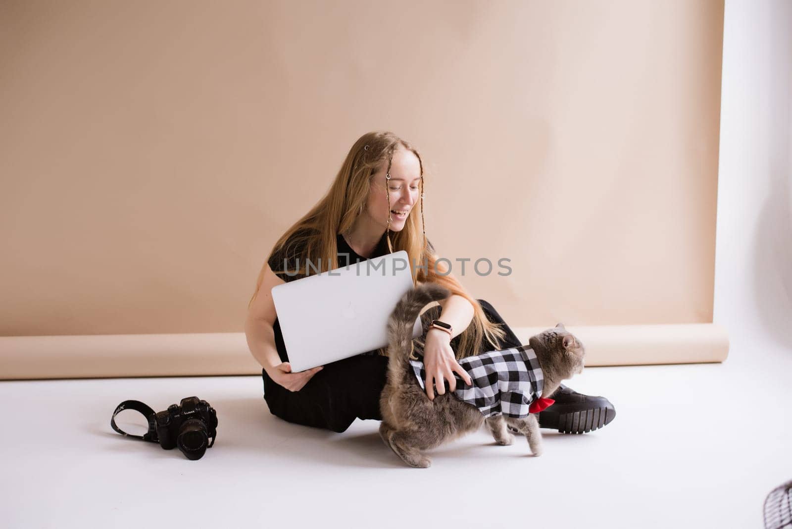 A business woman freelancer in a black is working, typing on a laptop apple and sitting on a white floor. blonde assistant in video production with computer MacBook and camera with cat