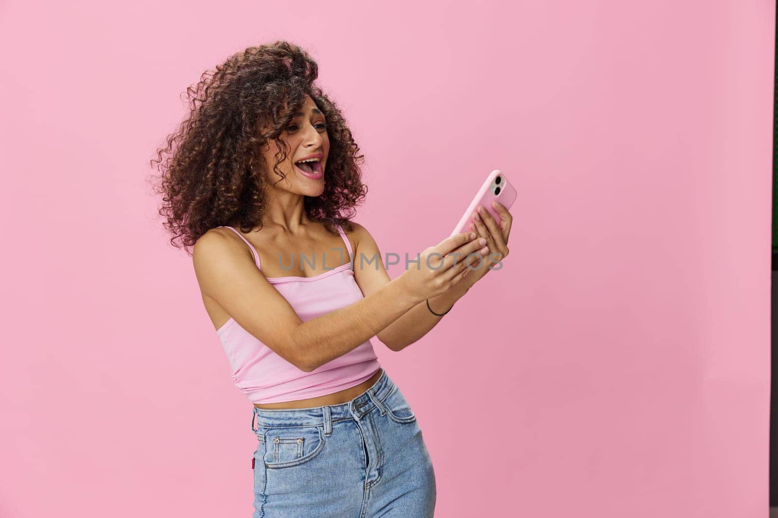 Woman blogger holding phone video call takes selfies, with curly hair in pink smile t-shirt and jeans poses on pink background, copy space, technology and social media, online by SHOTPRIME
