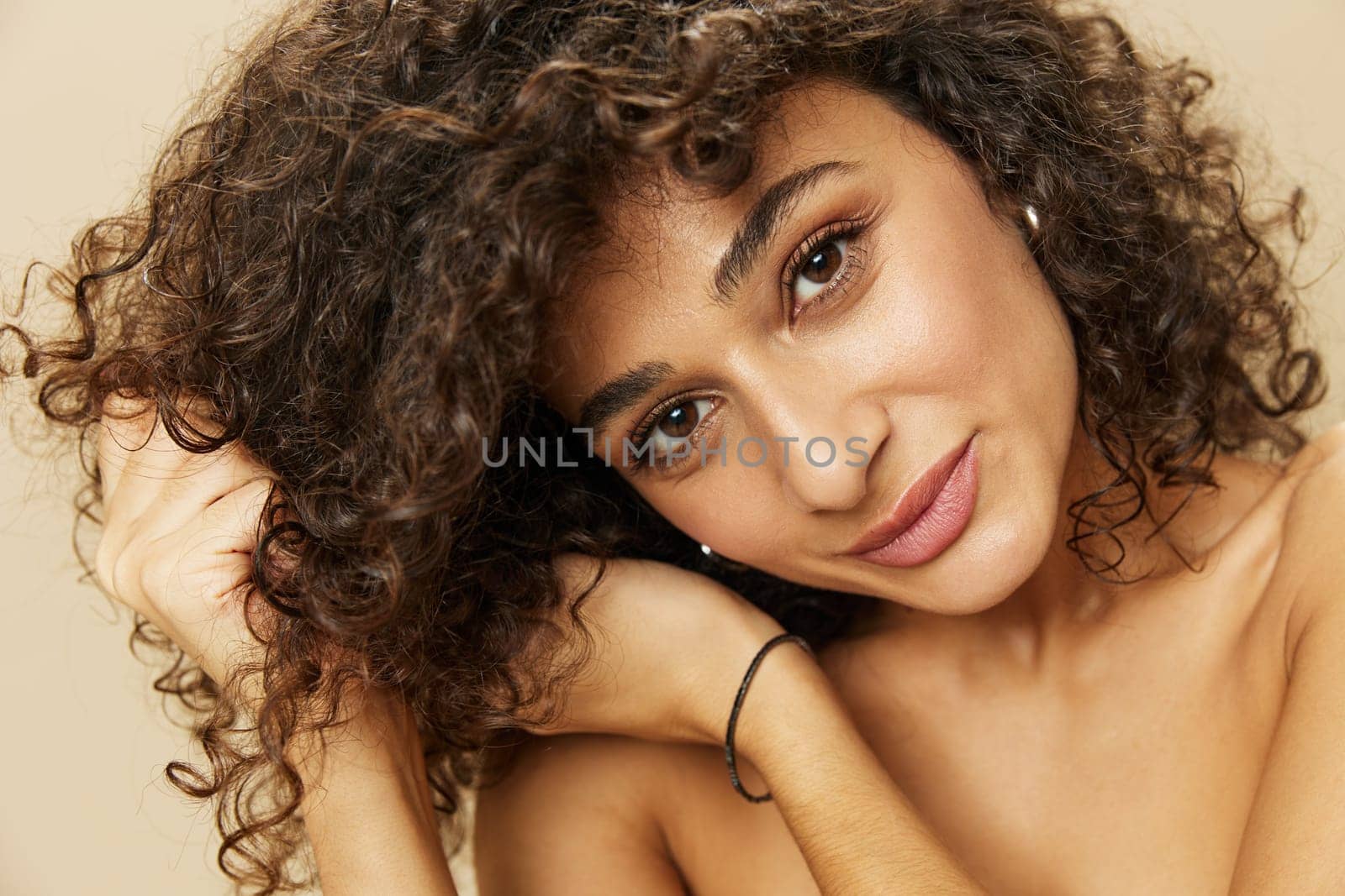 Woman applies cream and balm to her curly hair, the concept of protection and care, a healthy look, a smile with teeth on a beige background by SHOTPRIME