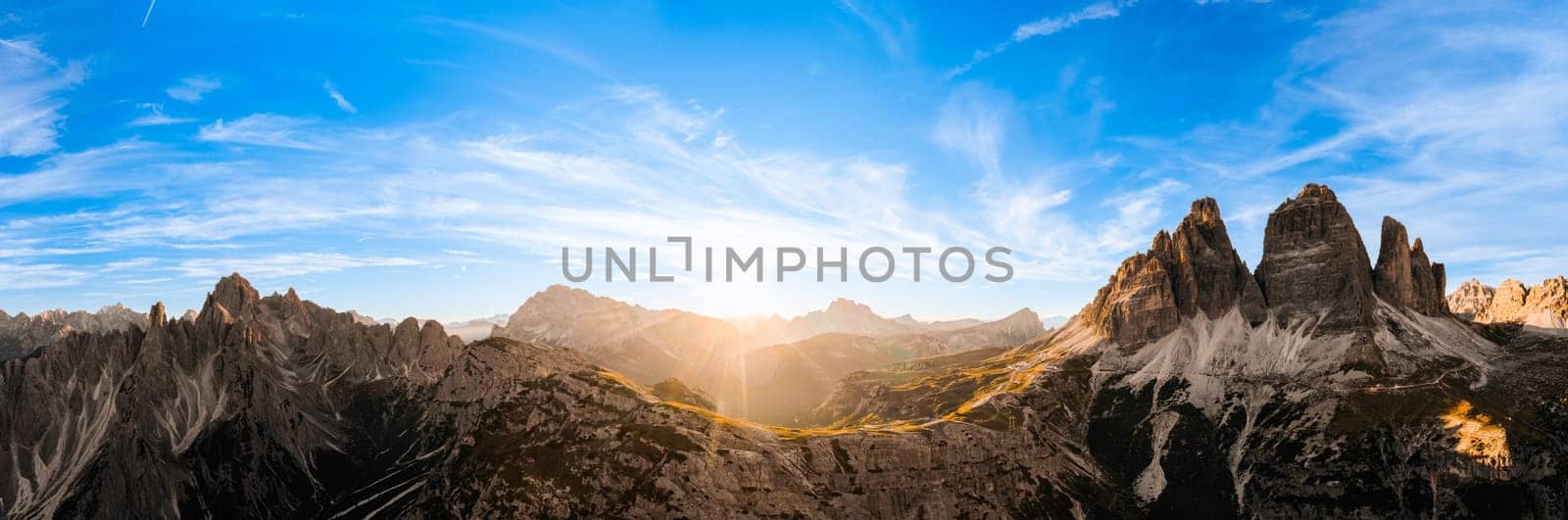 Sun sets on horizon above rocky mountain range. Sand-covered slopes of foothills of Tre Cime di Lavaredo silhouette at sunset aerial view in back lit