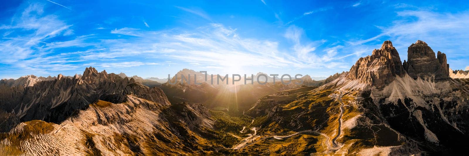 Curved road on rocky highland under blue sky with white clouds. Bare mountain landscape of Three Peaks of Lavaredo at sunset aerial view