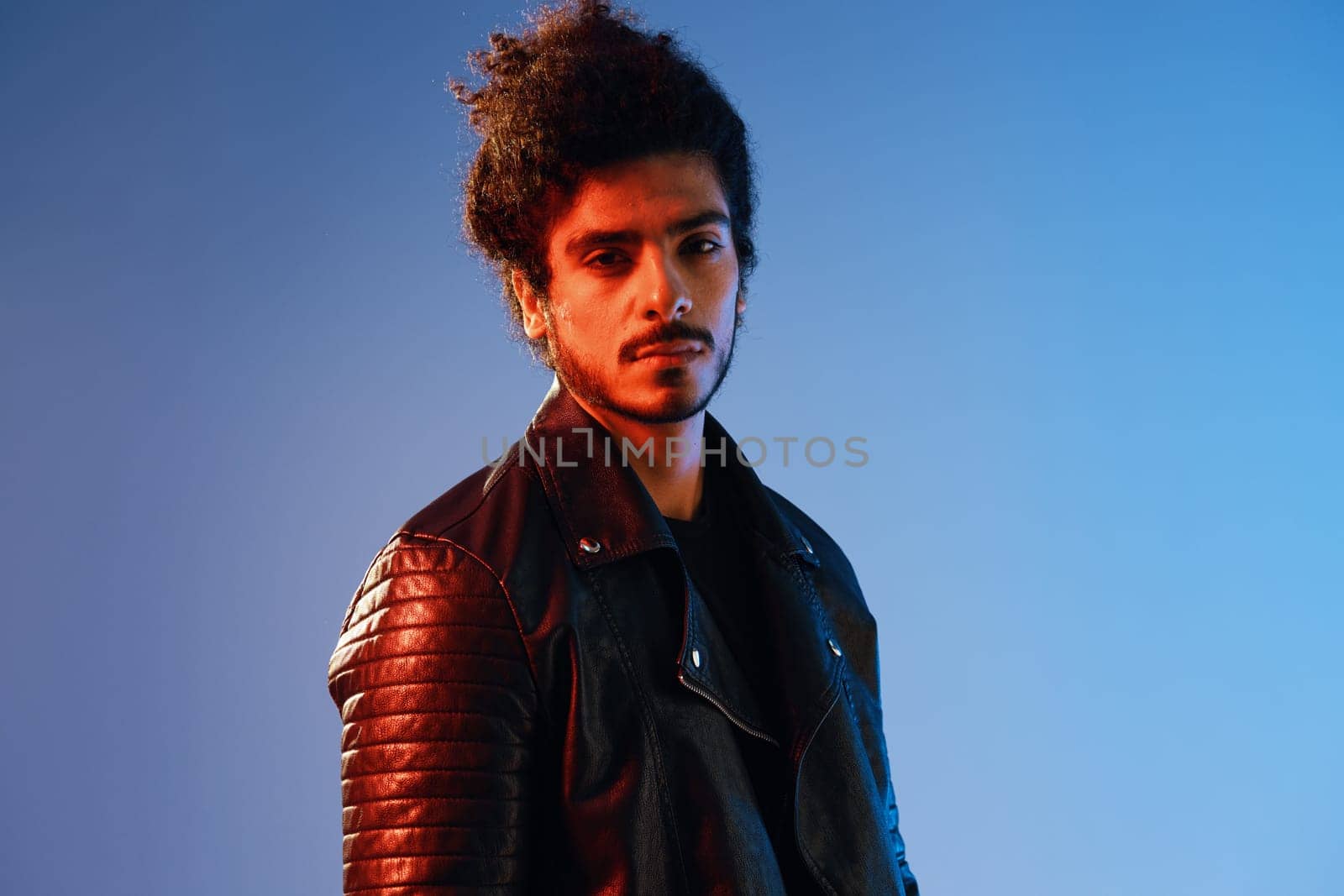 Portrait of a stylish man with curly hair on a blue background multinational, colored light, black leather jacket trend, modern concept. High quality photo