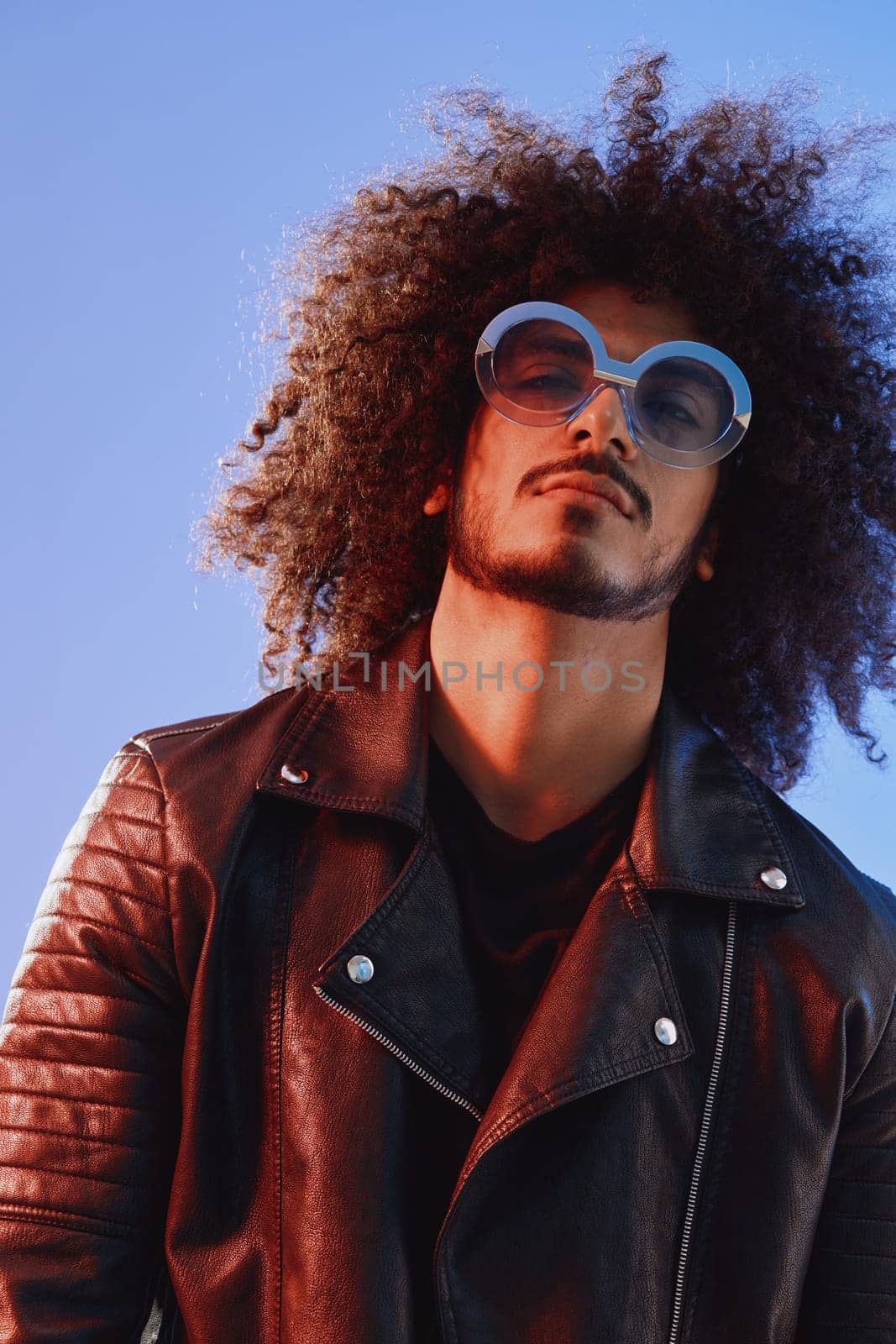 Portrait of a stylish man with curly hair with glasses smile on blue background multinational, color light, black leather jacket trend, modern concept. High quality photo