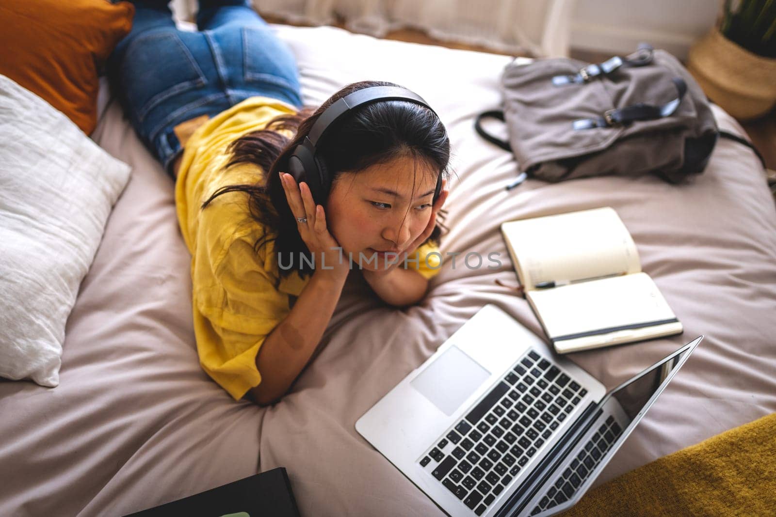 Asian female college student at home bedroom lying down on bed using laptop attending online university classes listening teacher lesson with headphones. E-learning concept.