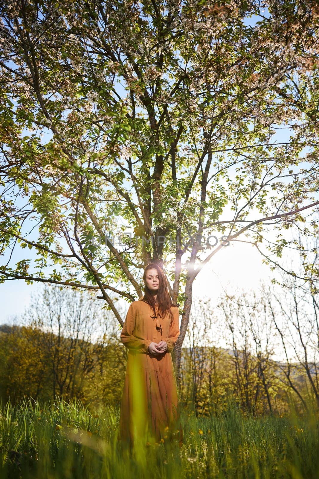 a sweet, modest woman with long hair stands in the countryside near a flowering tree and looks at the camera with her hands clasped together by Vichizh