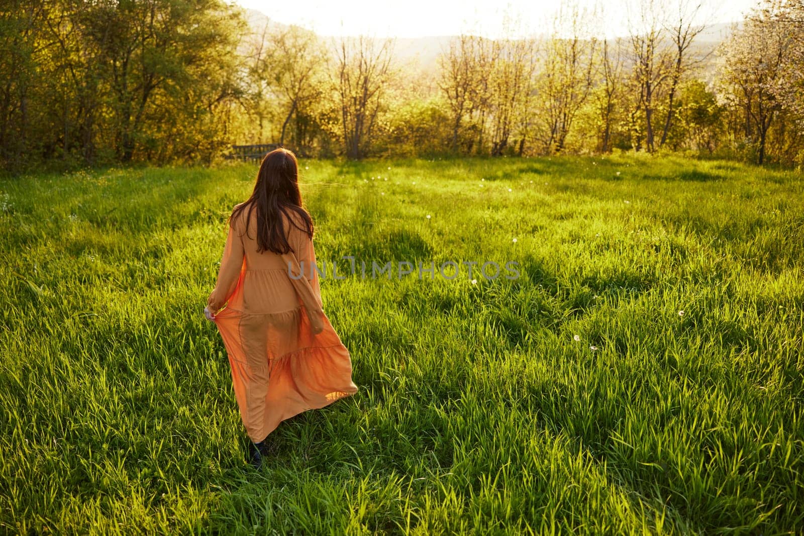 an attractive, slender, red-haired woman walks through a field during sunset, in a long orange dress enjoying unity with nature and relaxation holding the edge of her clothes by Vichizh