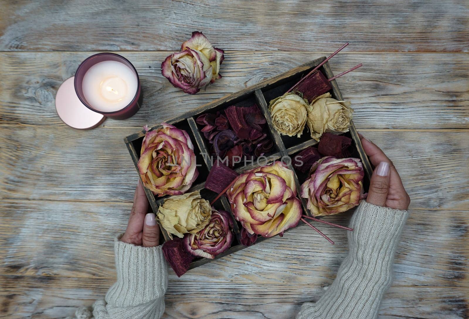 Women's hands are holding a box with dried rose flowers, there is a burning candle on the table. by Spirina