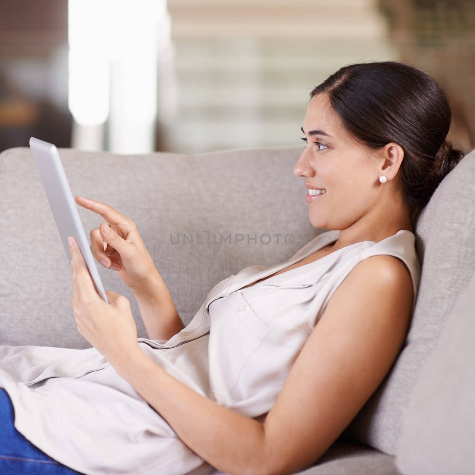 Happy woman, tablet and relax on living room sofa for streaming, browsing or social media at home. Female relaxing on technology for online entertainment, app or research lying on lounge couch by YuriArcurs