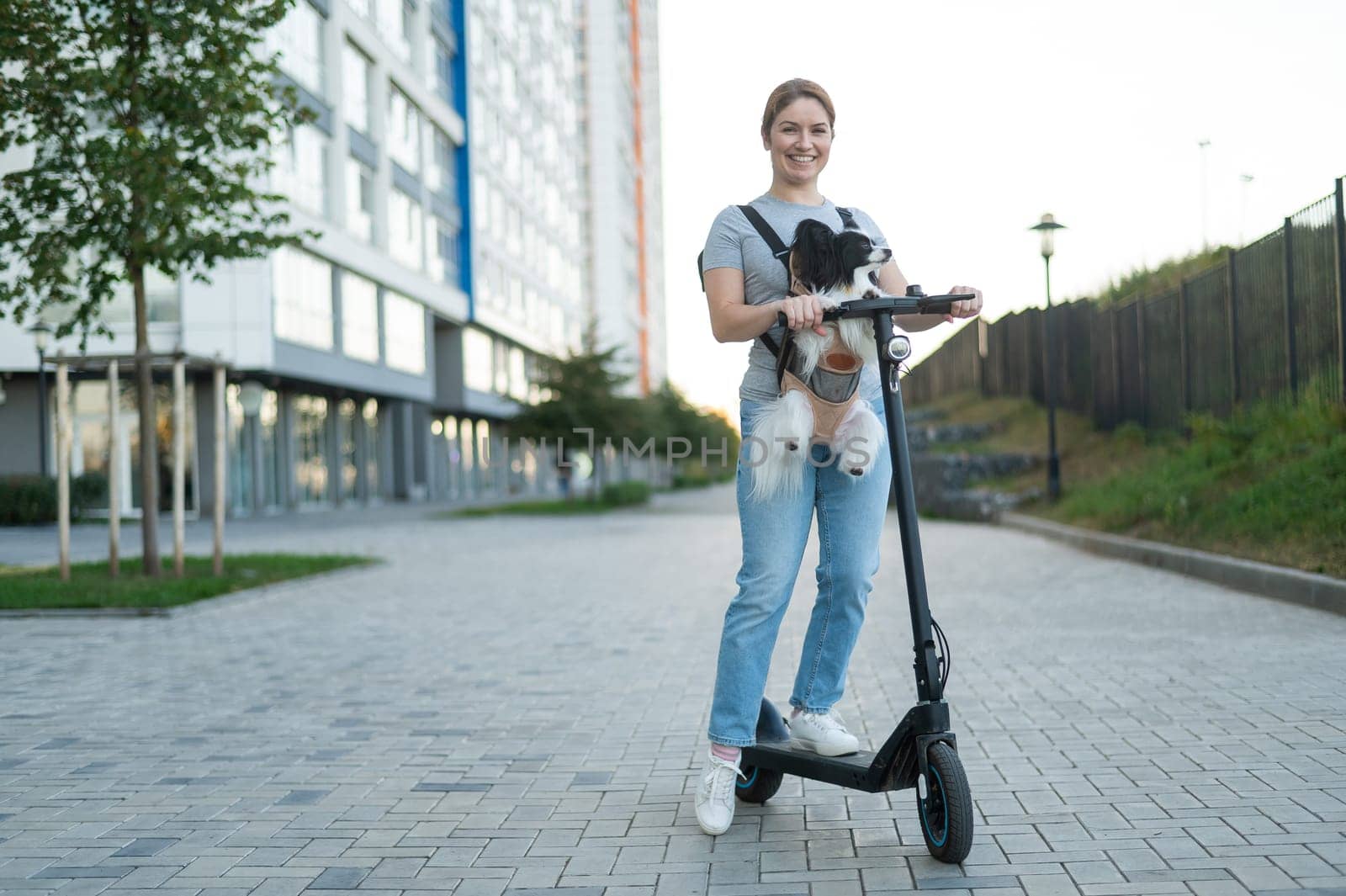A woman rides an electric scooter with a dog in a backpack. Pappilion Spaniel Continental in a sling