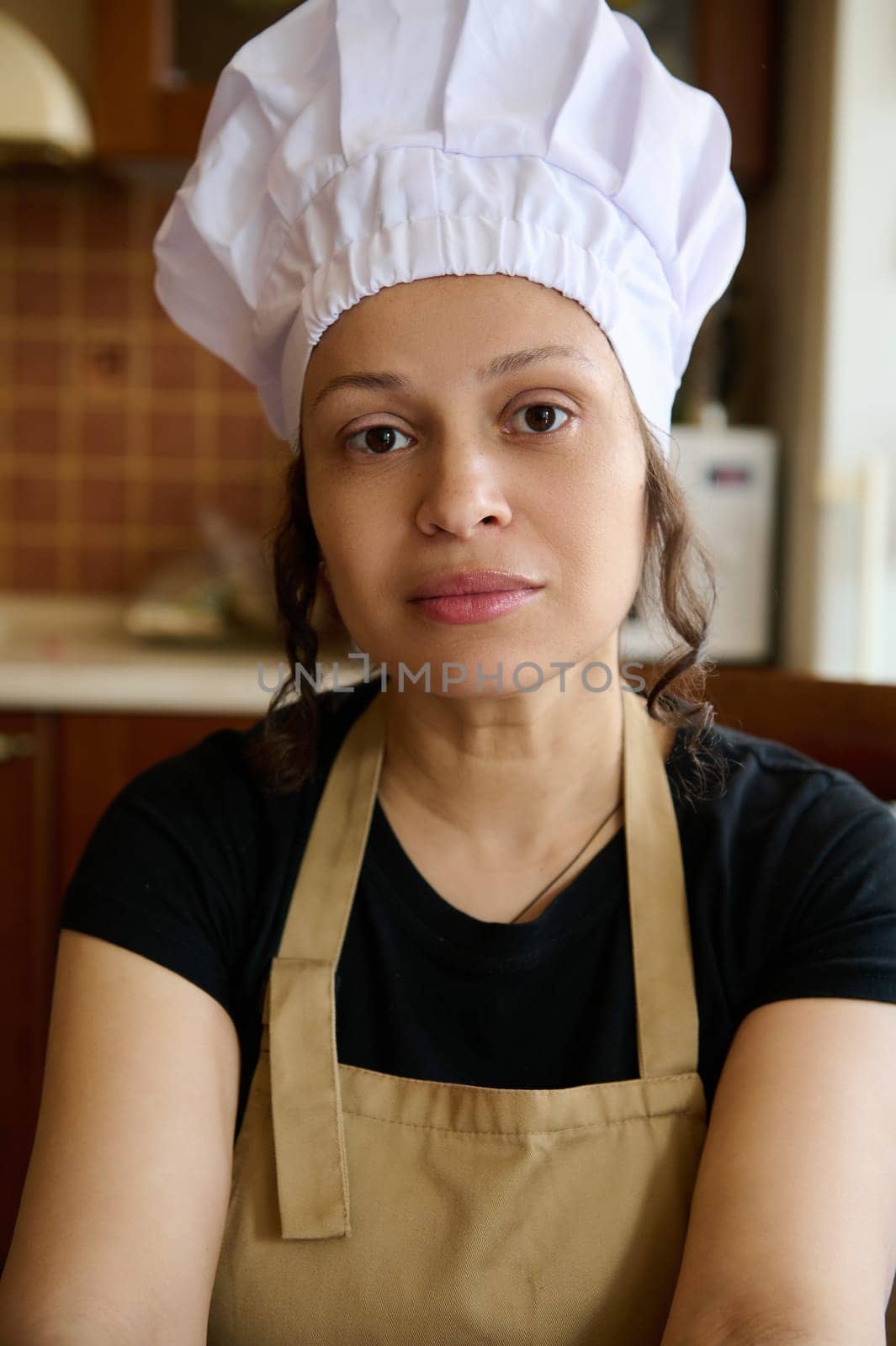 Confident portrait of a beautiful female baker confectioner, wearing white chef's hat and beige apron, looking at camera by artgf