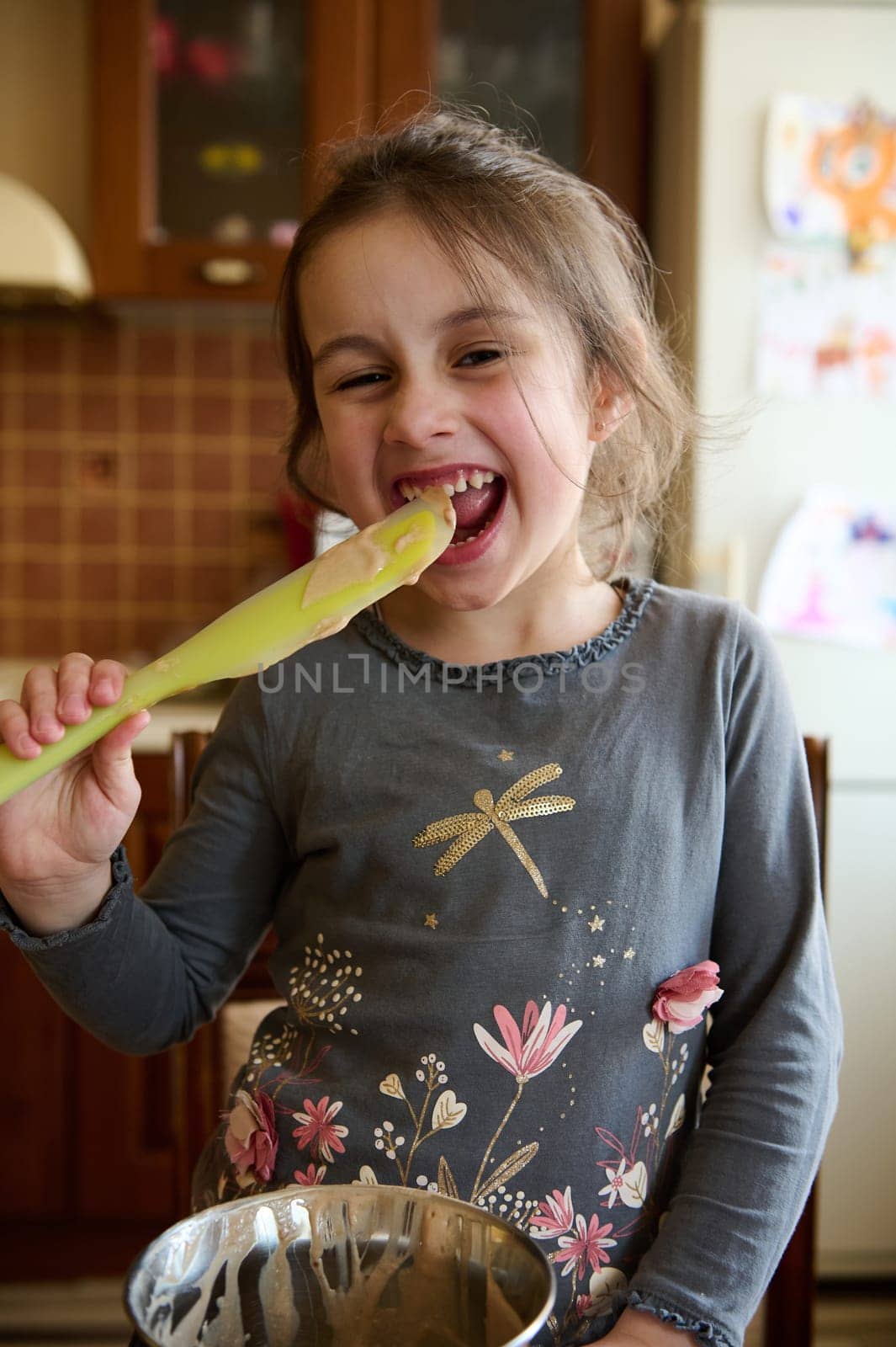 Cute little girl helps her mom in kitchen to prepare cream for the cake, quietly licks a spoon with melted delicious confectionery chocolate, mischievously smiling and making faces looking at camera