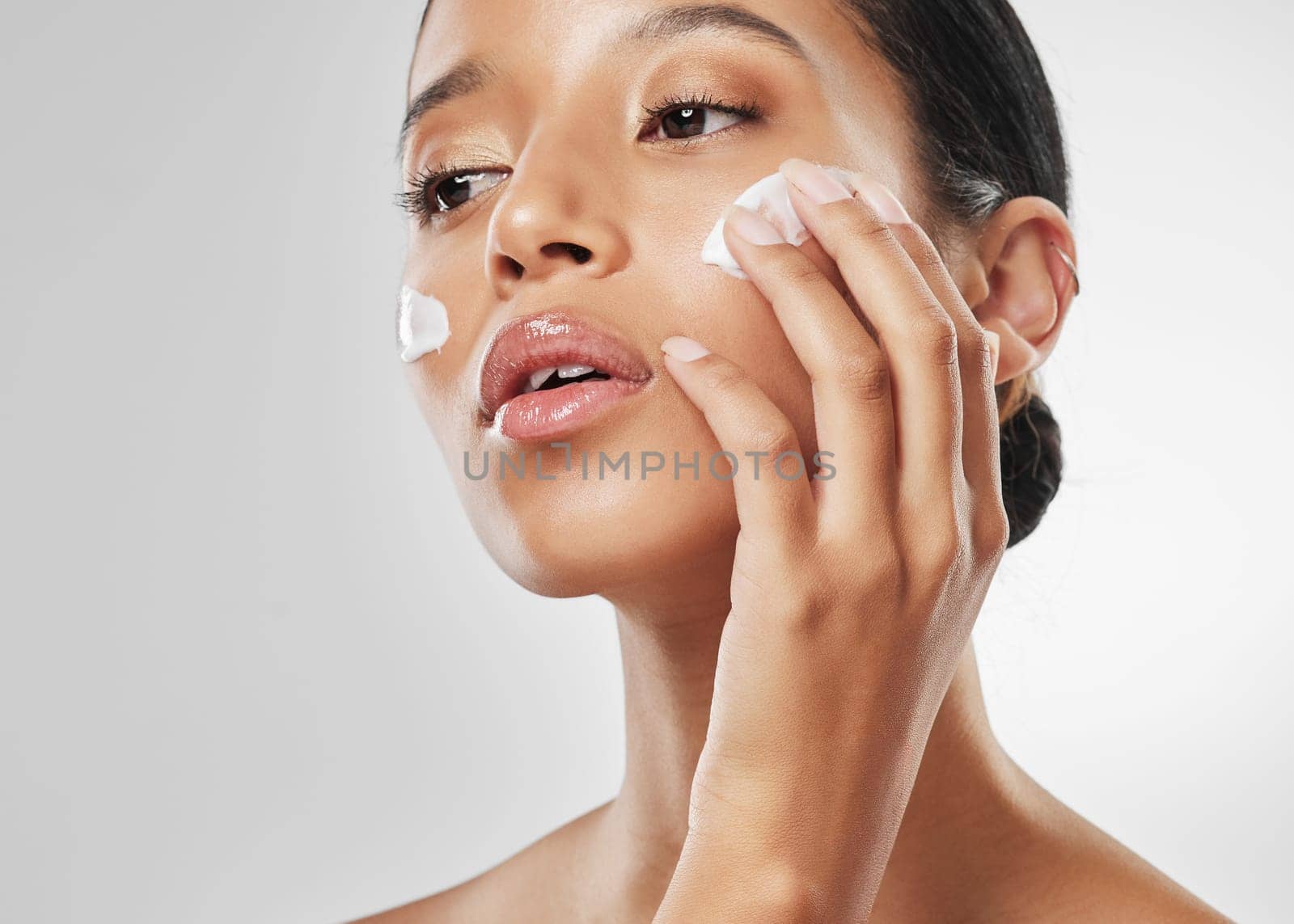 Refining texture and tone. Studio shot of an attractive young woman applying moisturiser on her face against a grey background. by YuriArcurs