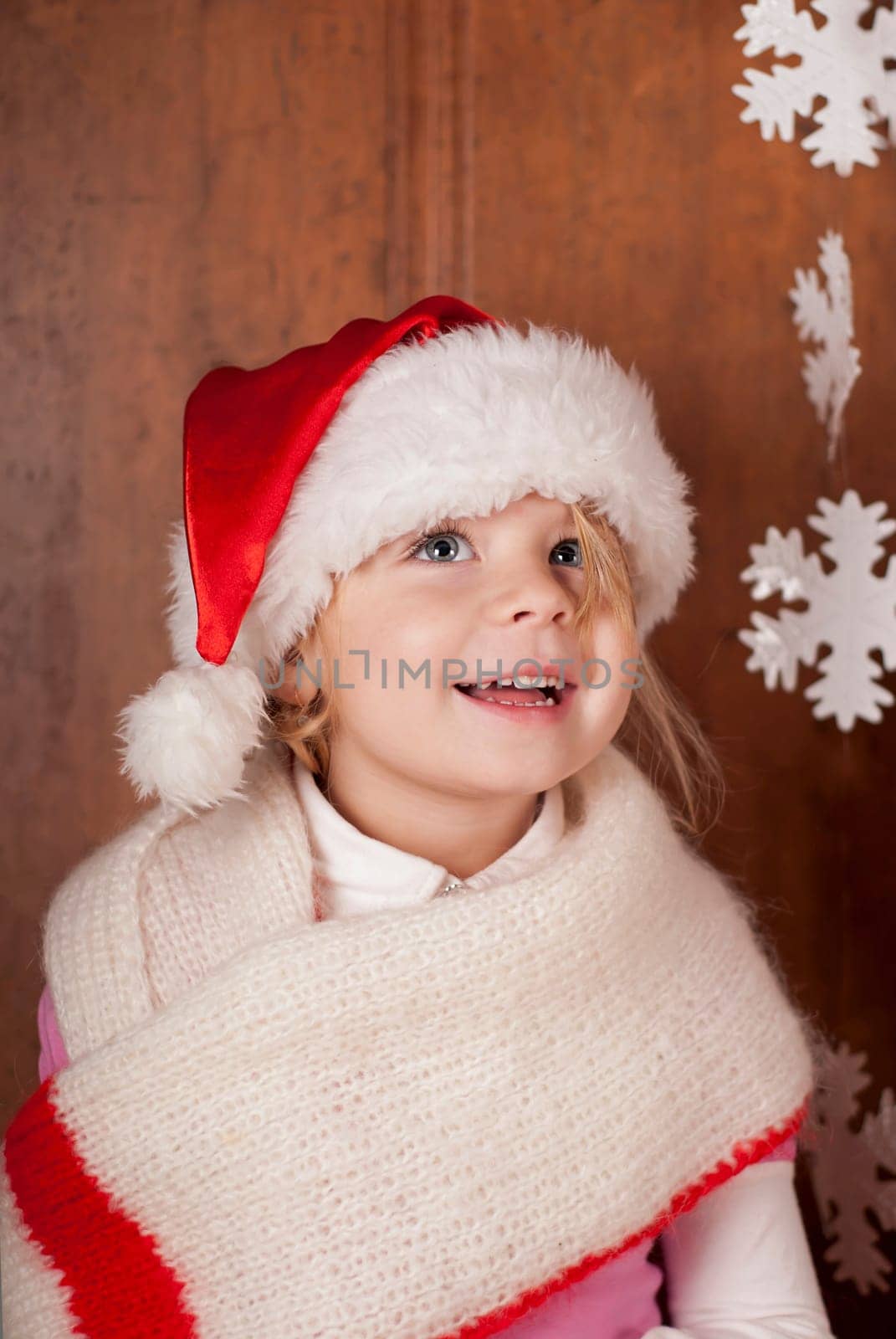 A girl in a warm red scarf and a santa claus hat examines paper snowflakes. The child rejoices in winter, New Year cartoons and the holiday of Christmas by aprilphoto