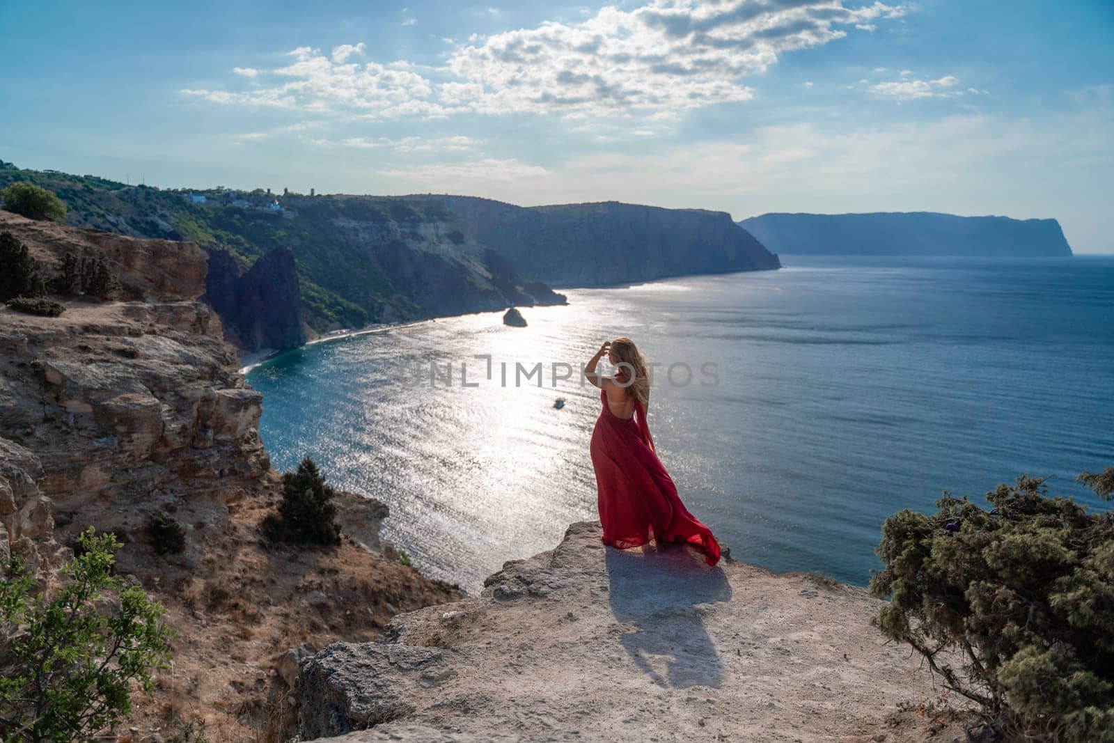 A girl with flowing hair in a long red dress stands on a rock above the sea. The stone can be seen in the sea. Sunny path to the sea from the sun. by Matiunina