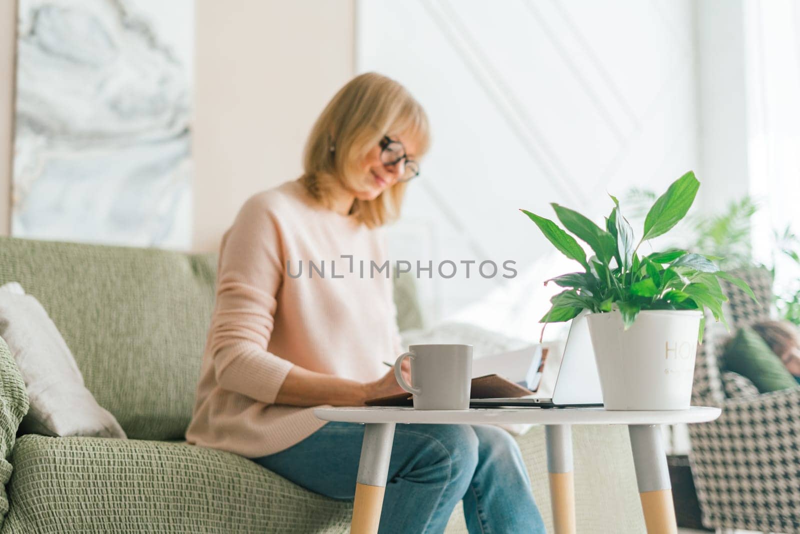 Cute adult senior mature woman in glasses working in her laptop computer. Beautiful seasoned female doing online banking investment trading in her laptop computer and drinking coffee from the mug.