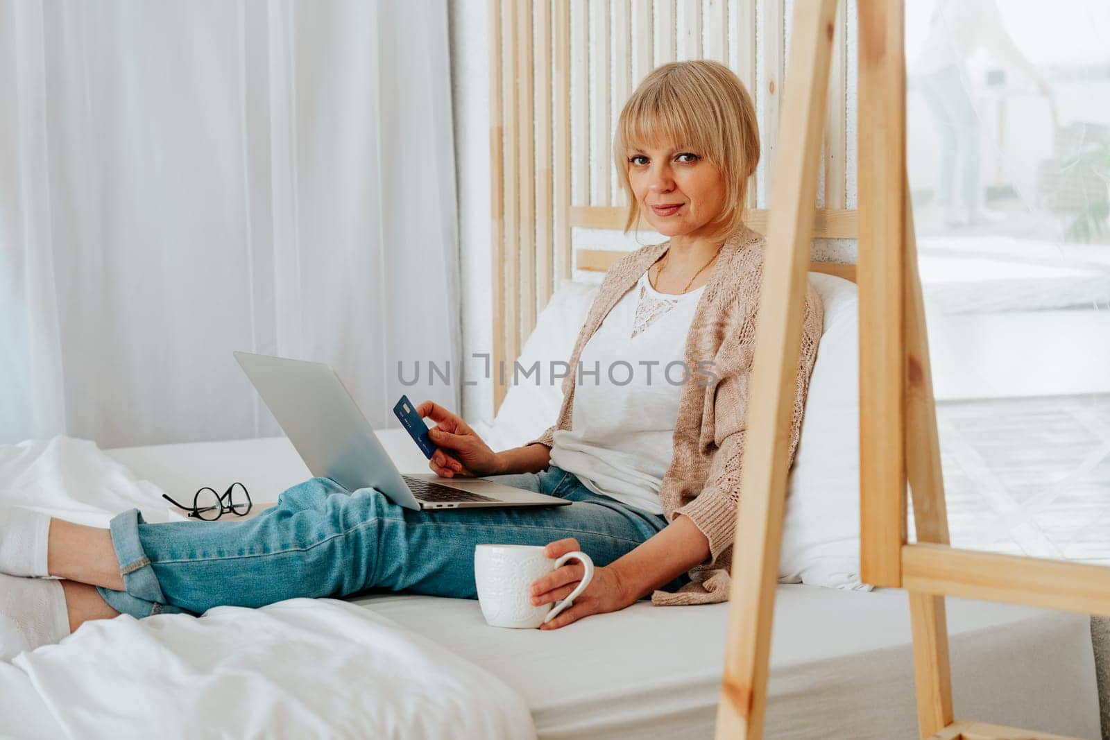 Senior mature woman sitting on bed with coffee tea mug and debit credit card while using her laptop for online shopping investments insurance by Ostanina