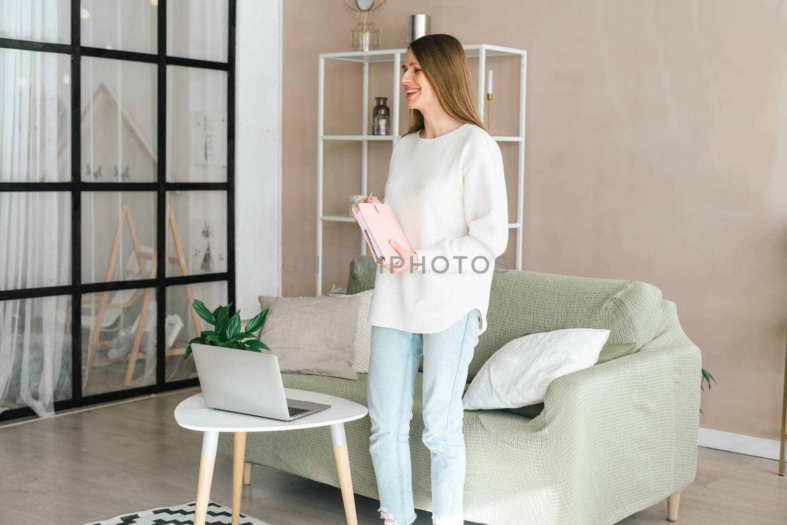 Smiling young woman girl standing near her laptop with the notepad notebook in her hands. Modern home office decor.