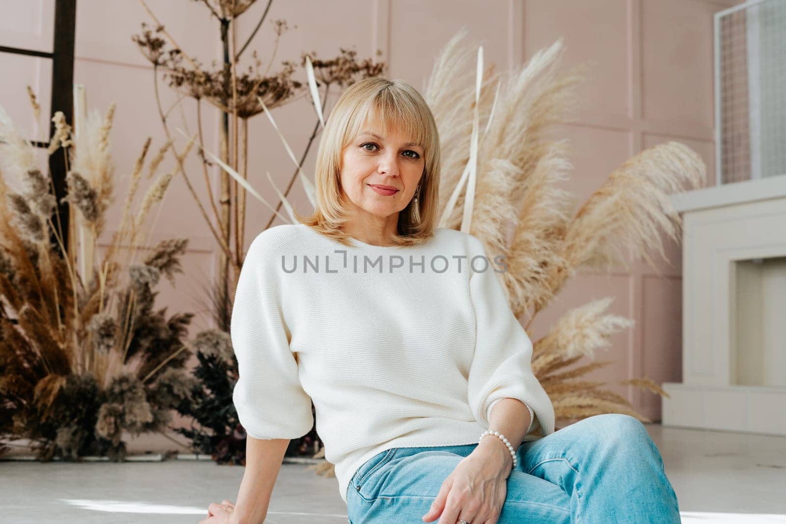 A portrait of cute senior mature adult woman in white sweater and blue jeans sitting on the floor with dry dried flowers in background by Ostanina
