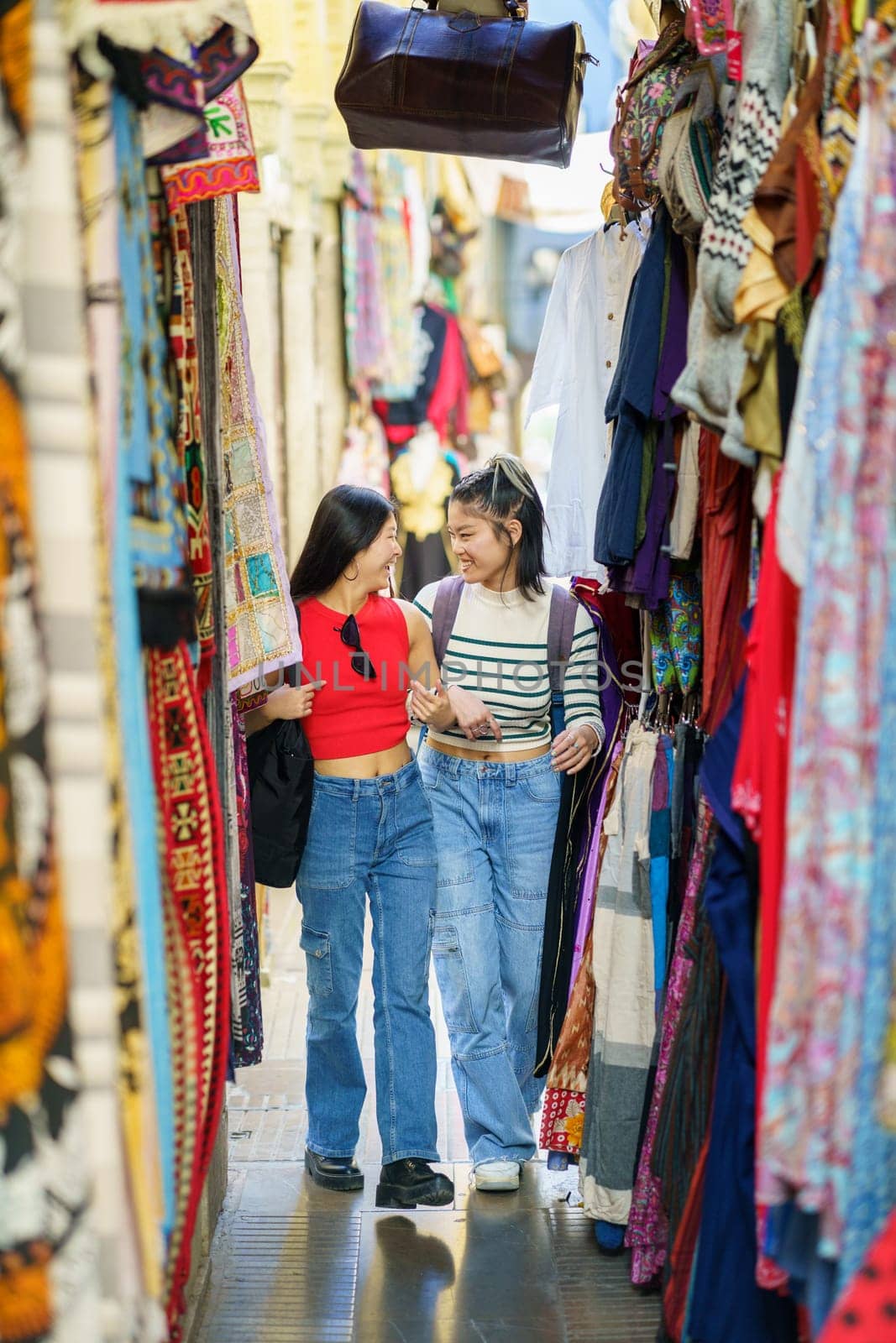 Smiling young women friends in casual clothes walking along colorful clothing store on street market in city during summer vacation