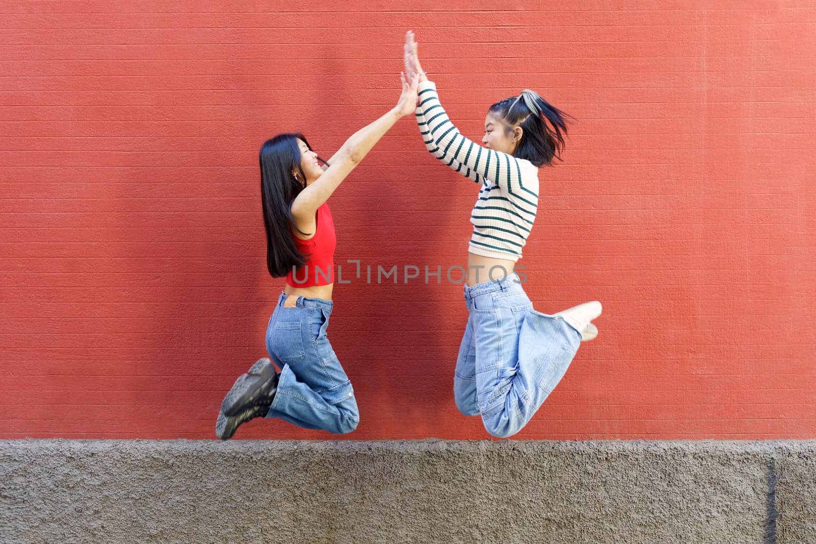 Full body side view of cheerful young Asian women friends jumping up and giving high five to each other against red wall