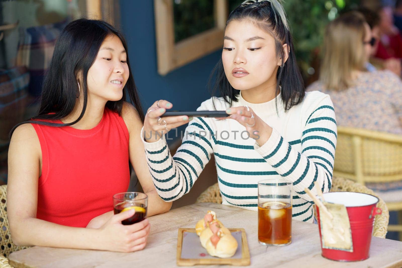 Smiling young Asian girl friends, in casual clothes sitting at table with cold drinks and taking picture of tasty food on smartphone while spending time together in cafe