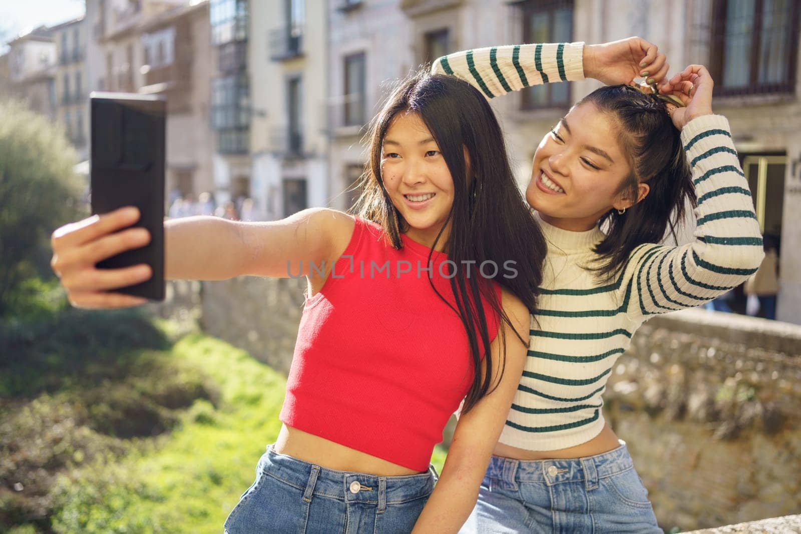 Young ethnic Asian girlfriends smiling happily while making selfie on cellphone together against city buildings in Granada