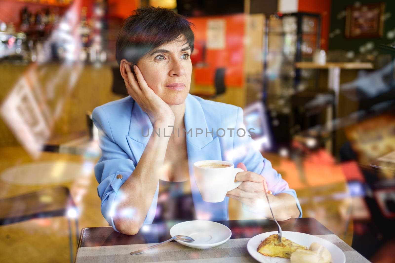 Pondering woman in stylish blue jacket sitting at table with cup of hot drink and looking away during breakfast in cafe