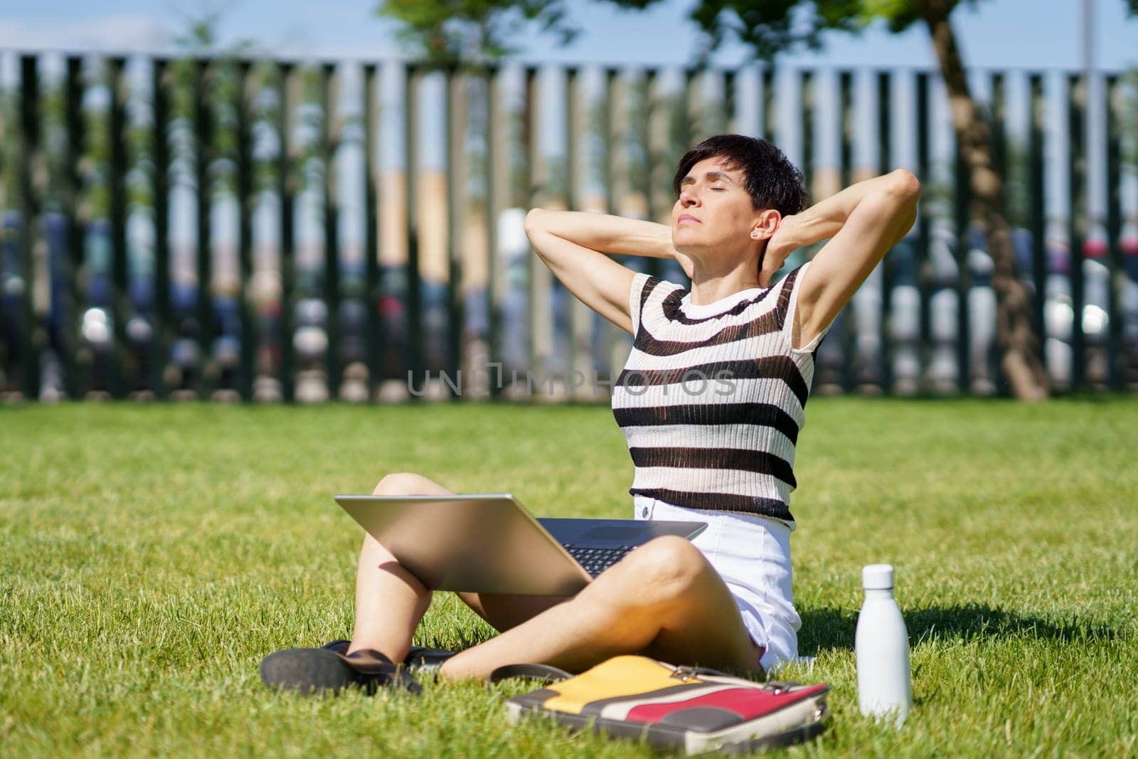 Full body of peaceful middle aged female freelancer, in summer clothes sitting on grass with laptop and closed eyes while stretching neck during remote work on project