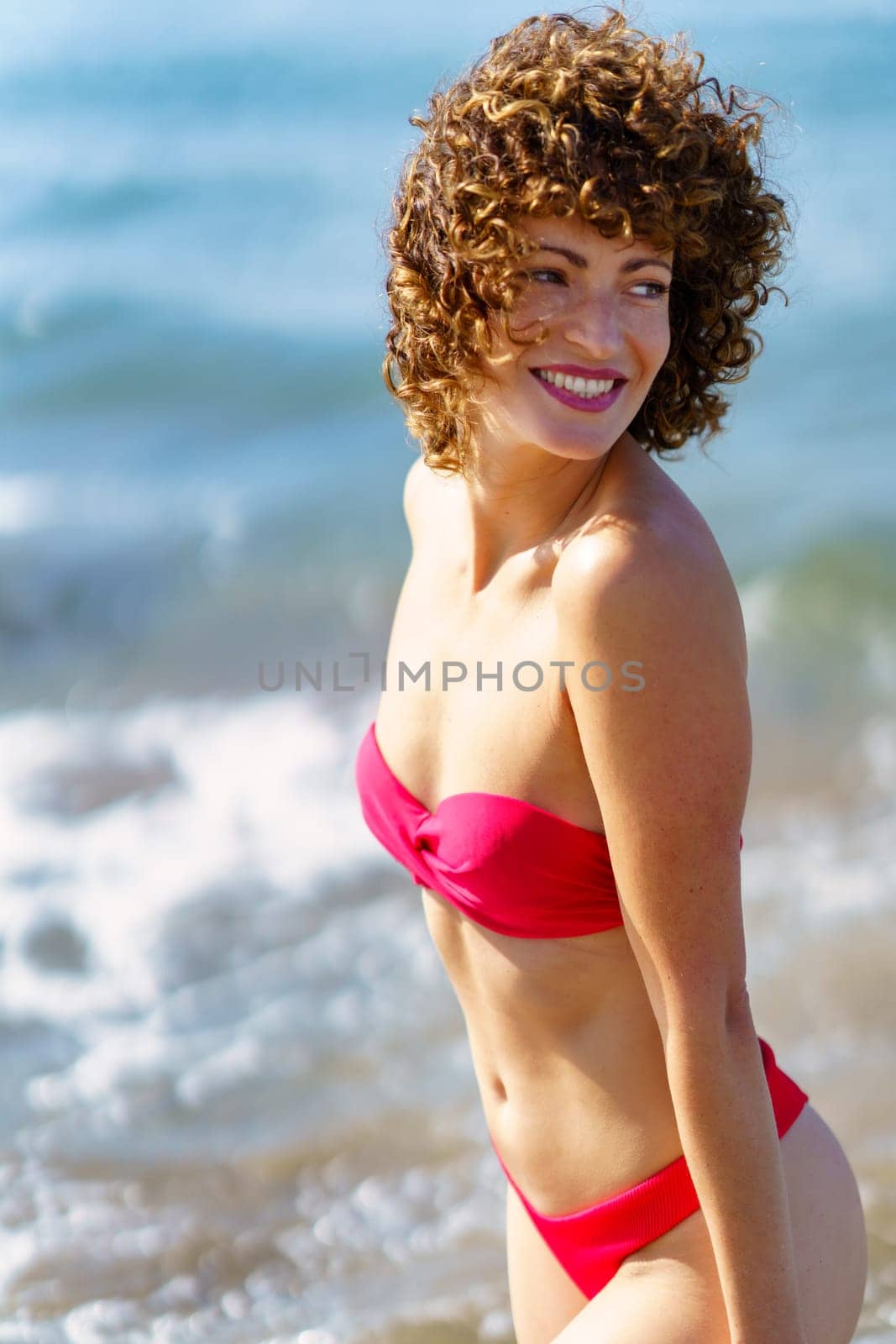 Side view of excited young female tourist with curly hair in stylish red bikini, smiling and looking over shoulder while standing on beach during summer vacation