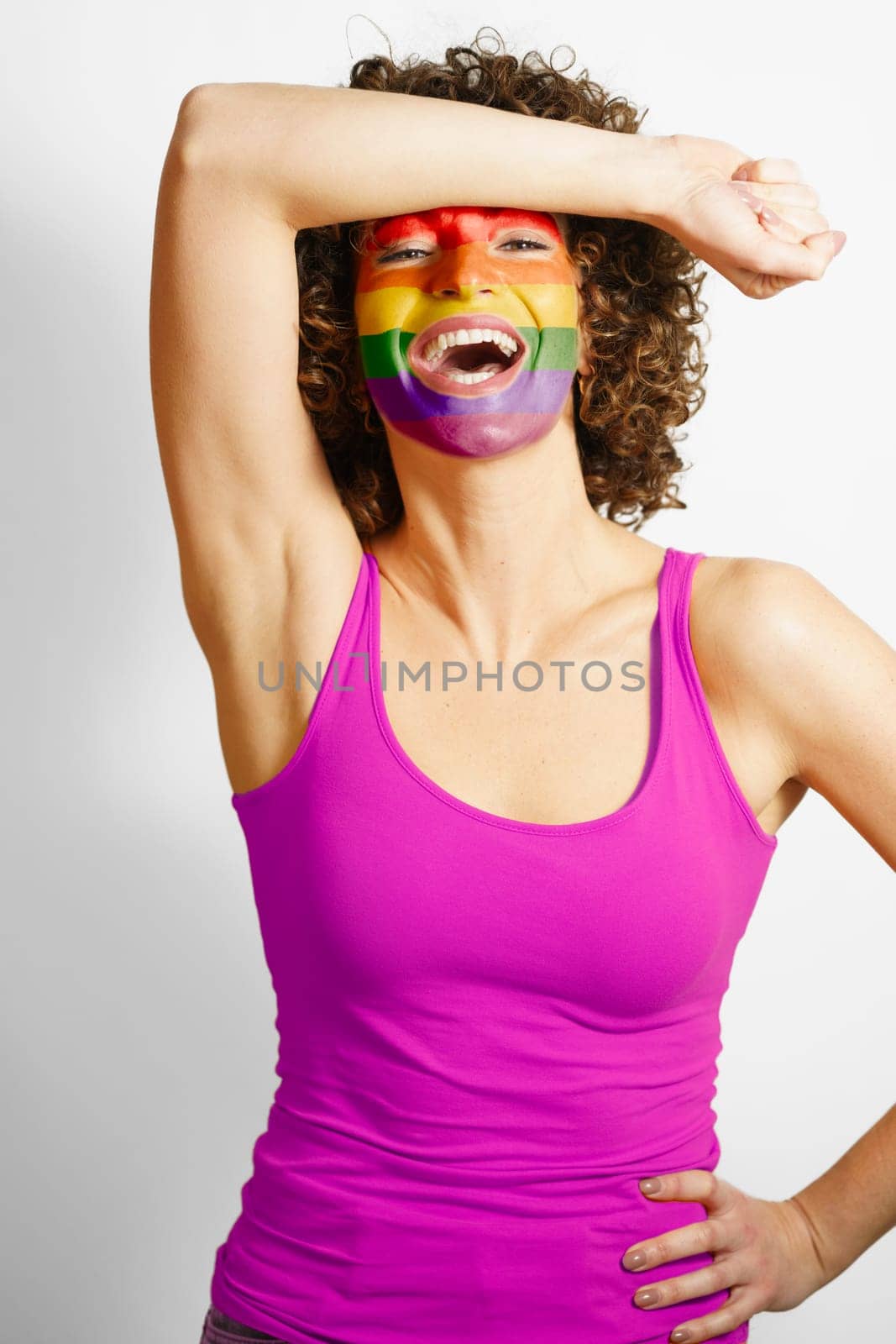 Cheerful female wearing purple tank top standing with hand on waist and painted in rainbow colors face looking at camera against white background