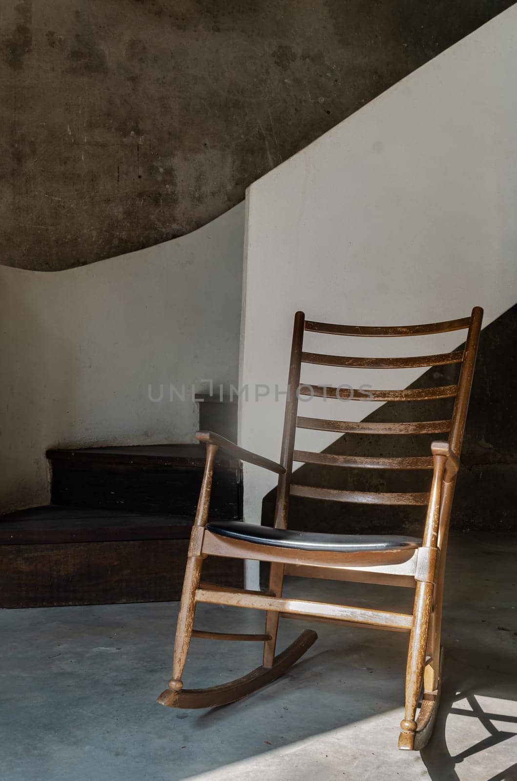 The Wooden Rocking Chair is standing alone in front of the White Spiral Staircase and Bare Cement Wall with Sunlight Shining Through. Space for text, Selective focus.