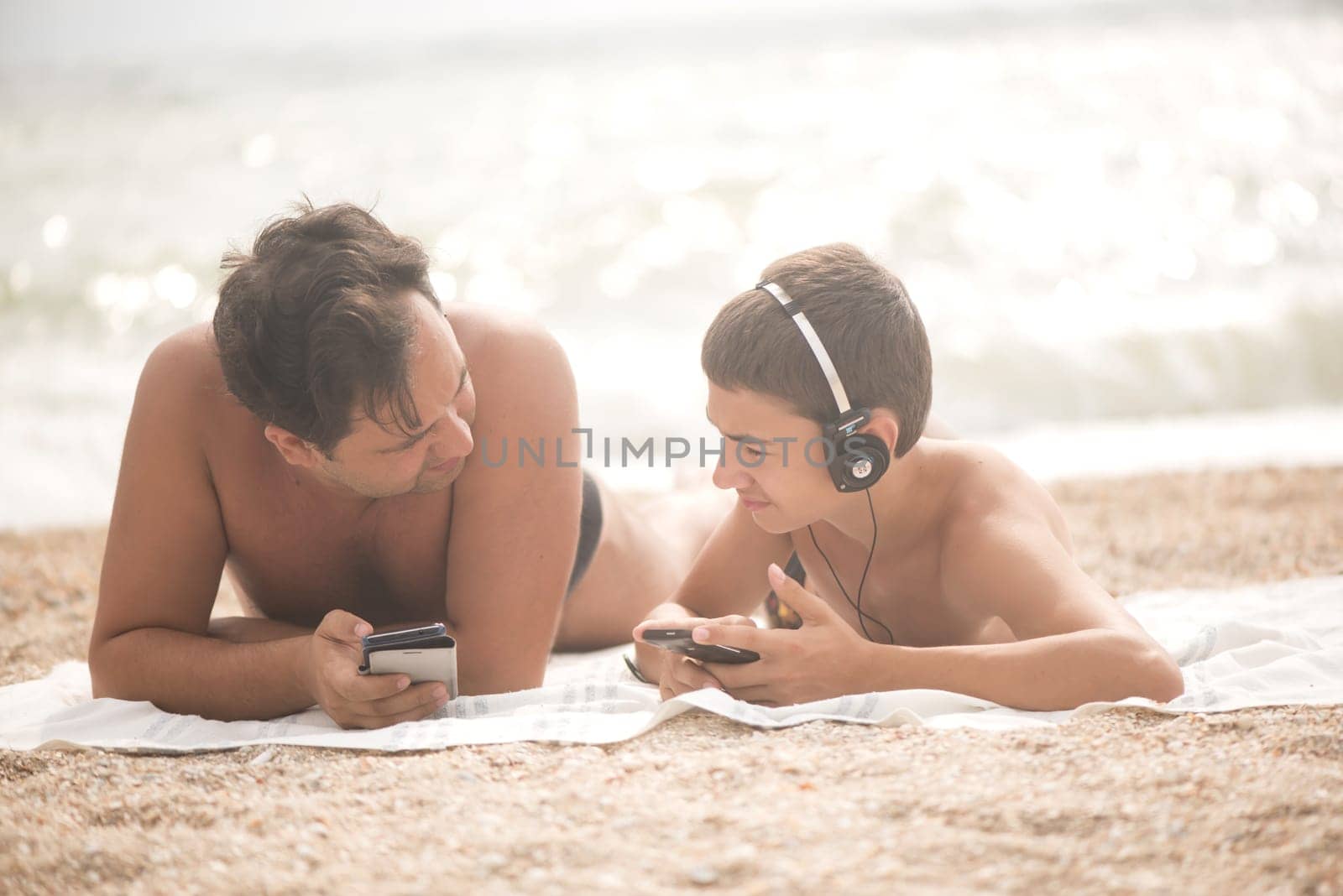 The sea is stormy. Sea of Azov. Teenager, boy in headphones with father with mobile devices in their hands communicate, read and listen to music on a beach on the sea by aprilphoto