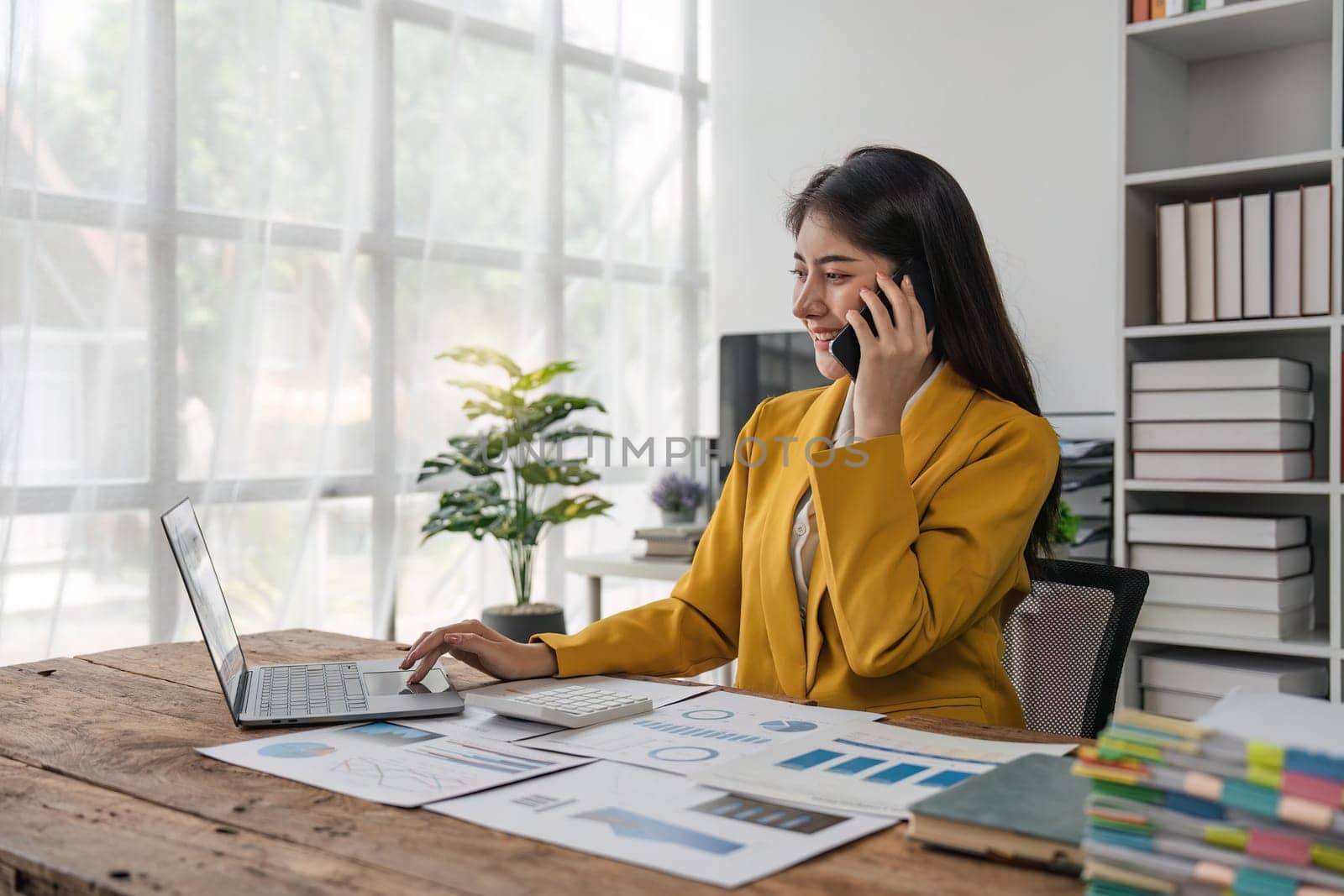 Portrait of financial business woman sitting at desk and working on laptop while making talk on the phone.