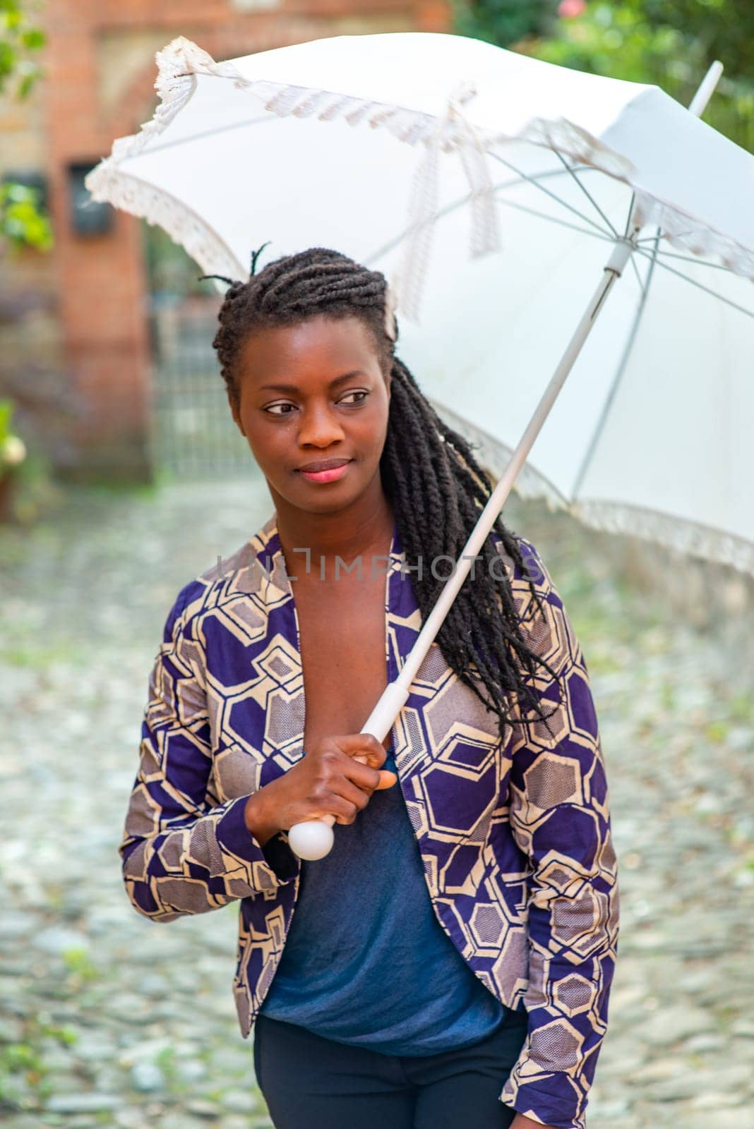 Young woman with dreadlocks braided hairstyle holding umbrella parasol. Happy young woman feeling confident in her style. Fashionable woman standing in the street of old village against stone wall.