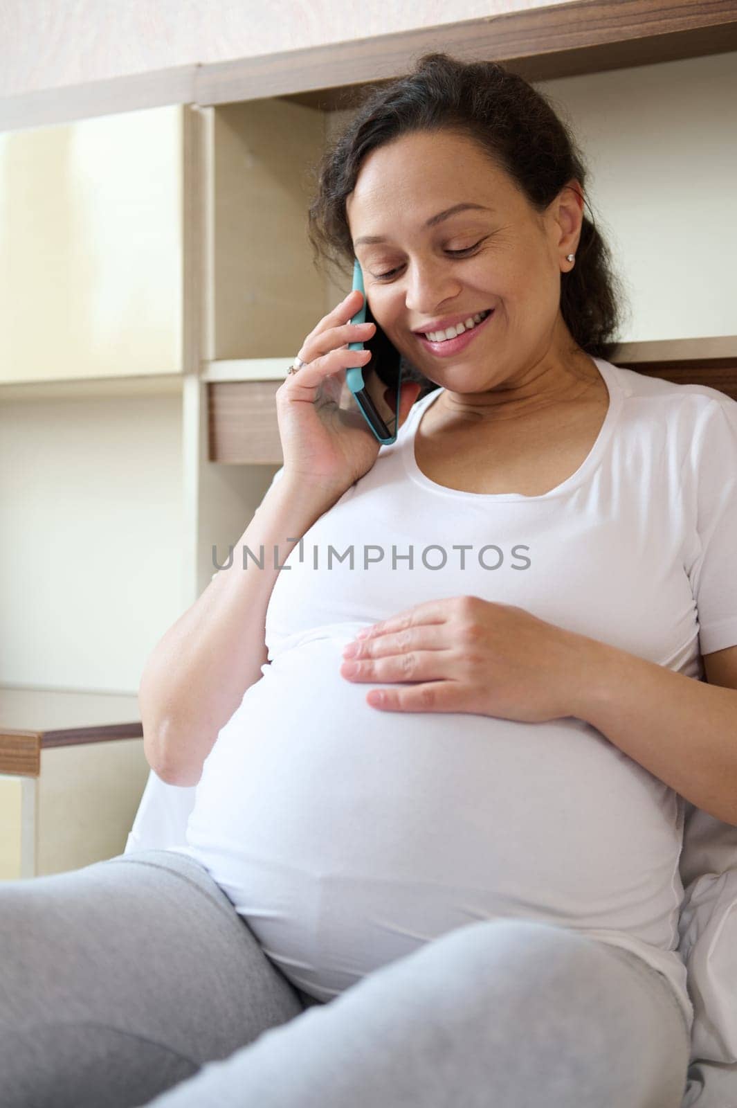 Happy smiling pregnant woman talking on mobile phone, touching her big belly while sitting relaxed on the bed at home by artgf