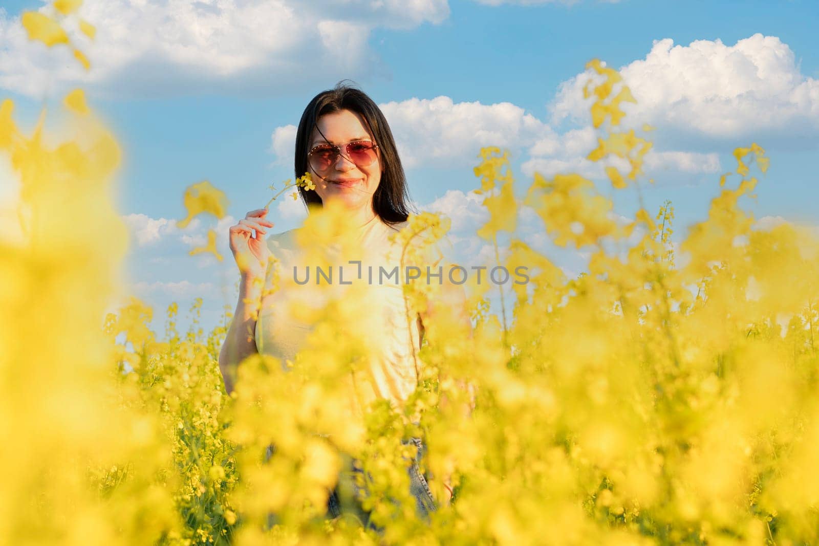 A beautiful girl in sunglasses stands in a field of yellow flowers against a blue sky, smiles happily and inhales the fresh spring and clean air. Peace concept. The color of the flag of Ukraine. No war.