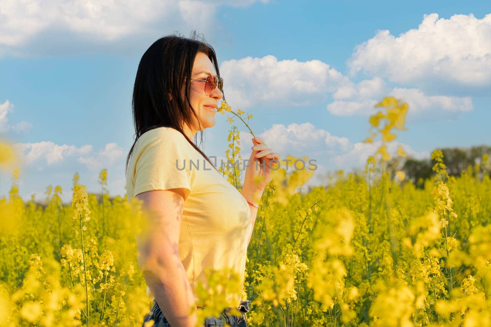 A beautiful girl in sunglasses stands in a field of yellow flowers against a blue sky, smiles happily and inhales the fresh spring and clean air. Peace concept. The color of the flag of Ukraine. No war.