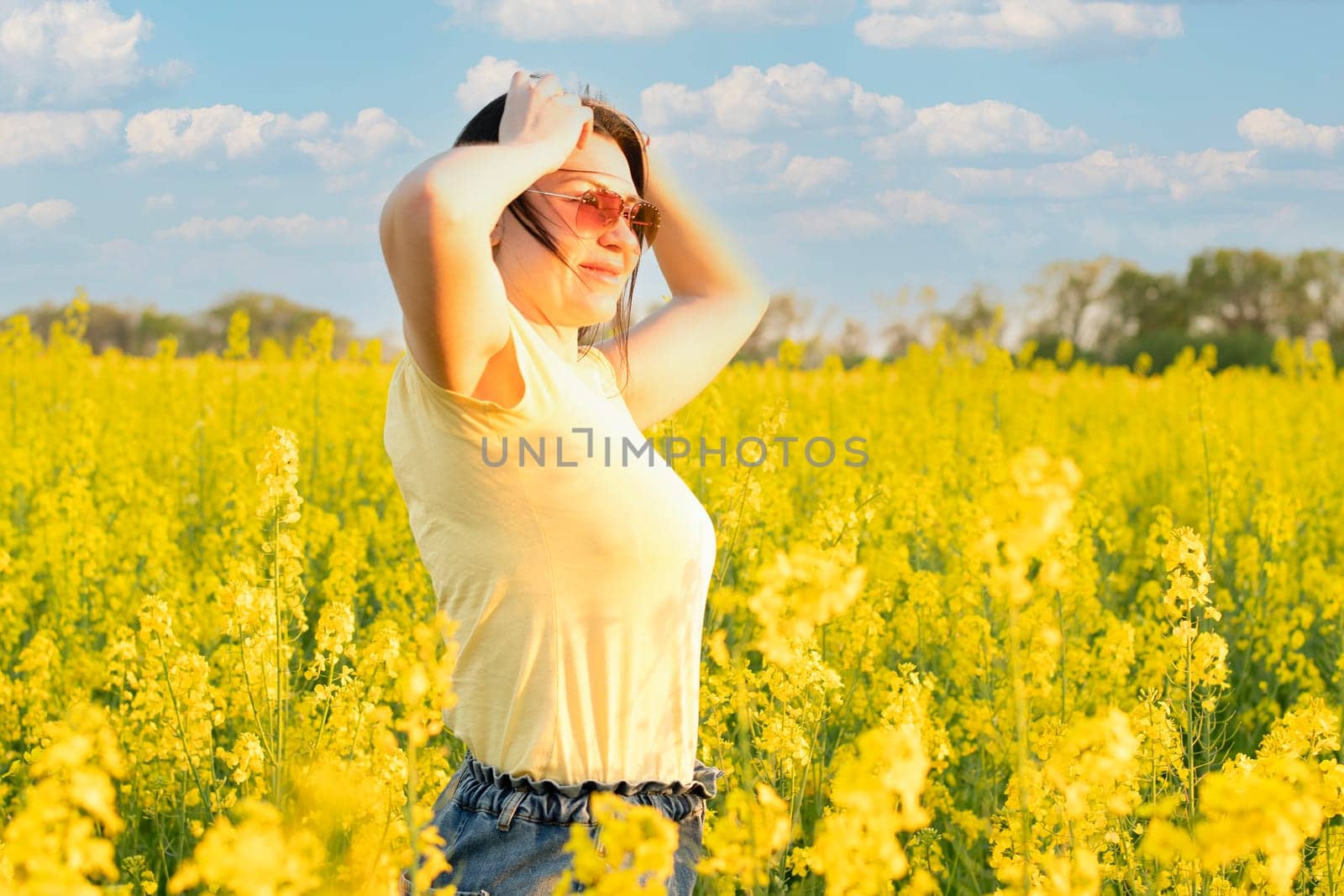 A beautiful girl in sunglasses stands in a field of yellow flowers against a blue sky, smiles happily and inhales the fresh spring and clean air. Peace concept. The color of the flag of Ukraine. No war.