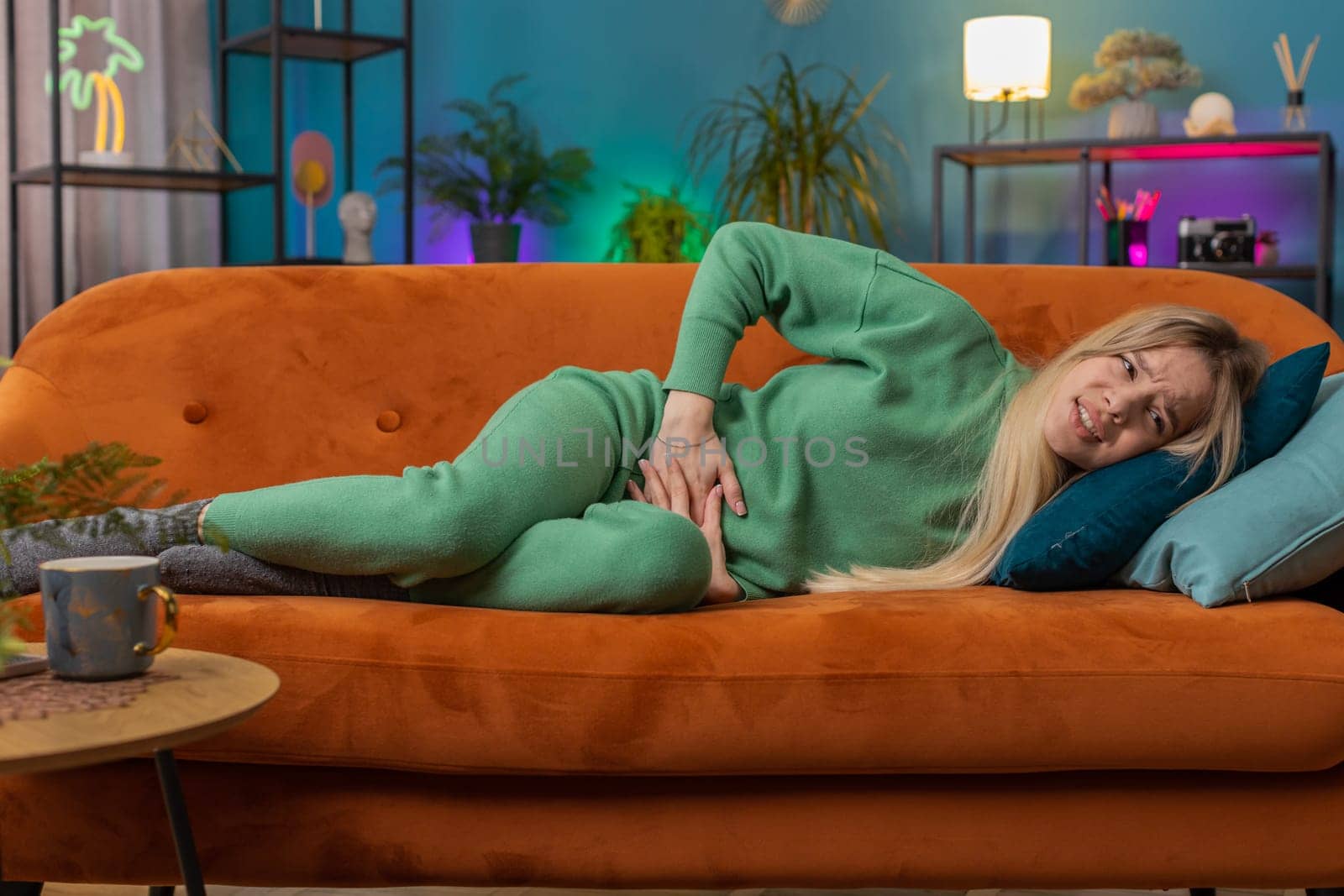 Sick ill woman suffering from period cramps, painful stomach ache lying on sofa at home room. Girl holding belly, feeling abdominal or menstrual pain. Abdominal pain, gastritis, diarrhea, indigestion