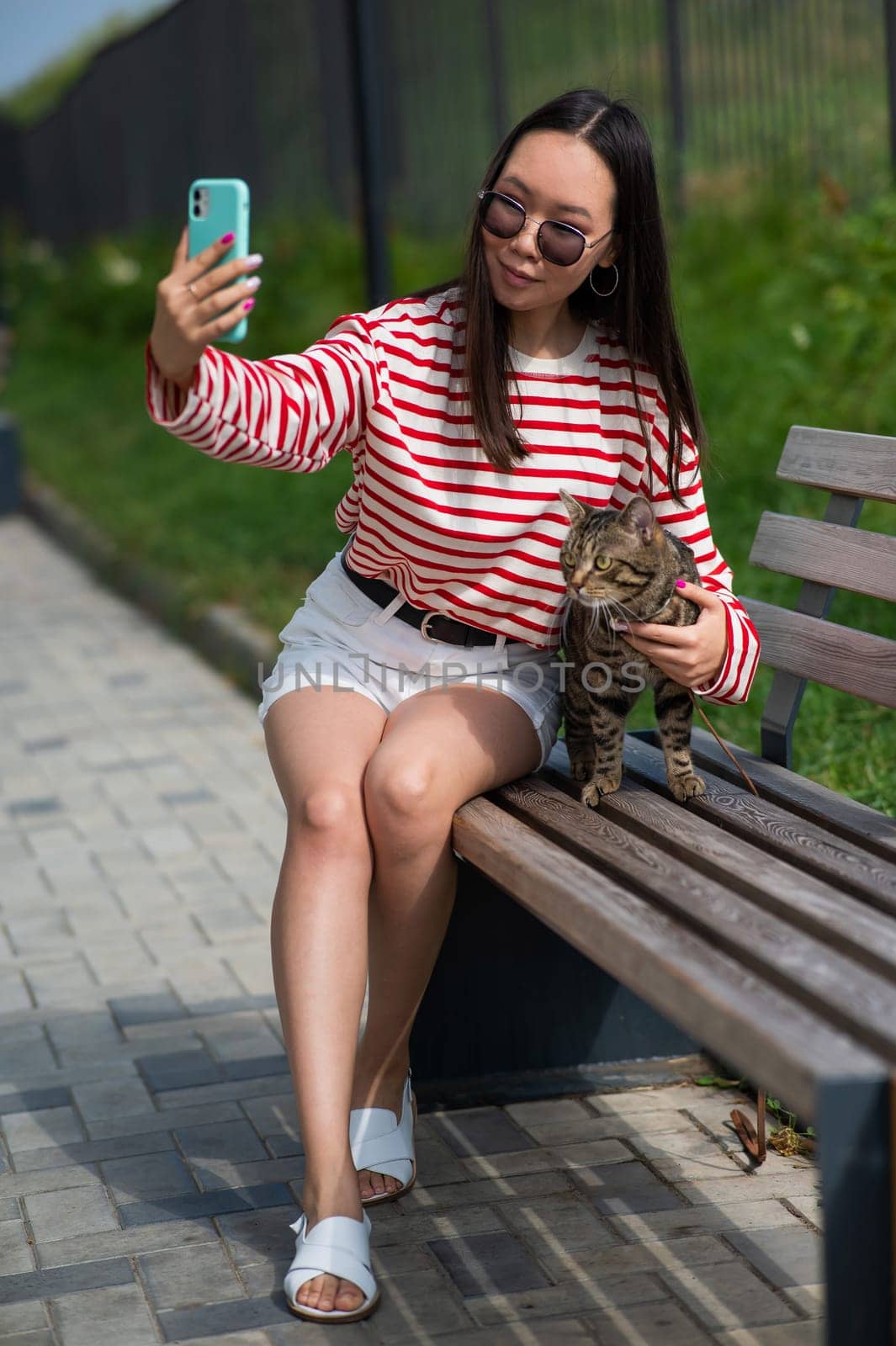 Young woman sits on a bench with a tabby cat and takes a selfie on a smartphone outdoors